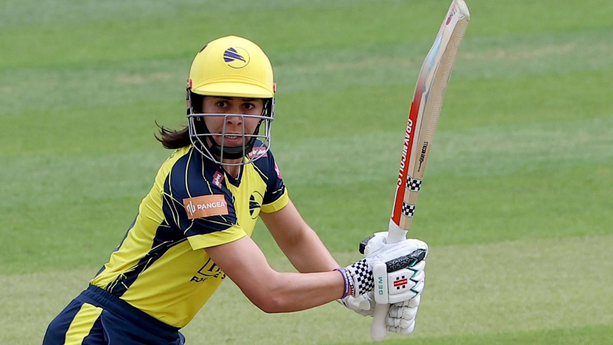 Hampshire's Maia Bouchier, wearing a yellow and blue kit and yellow cricket helmet, watches the ball to her right after playing a shot
