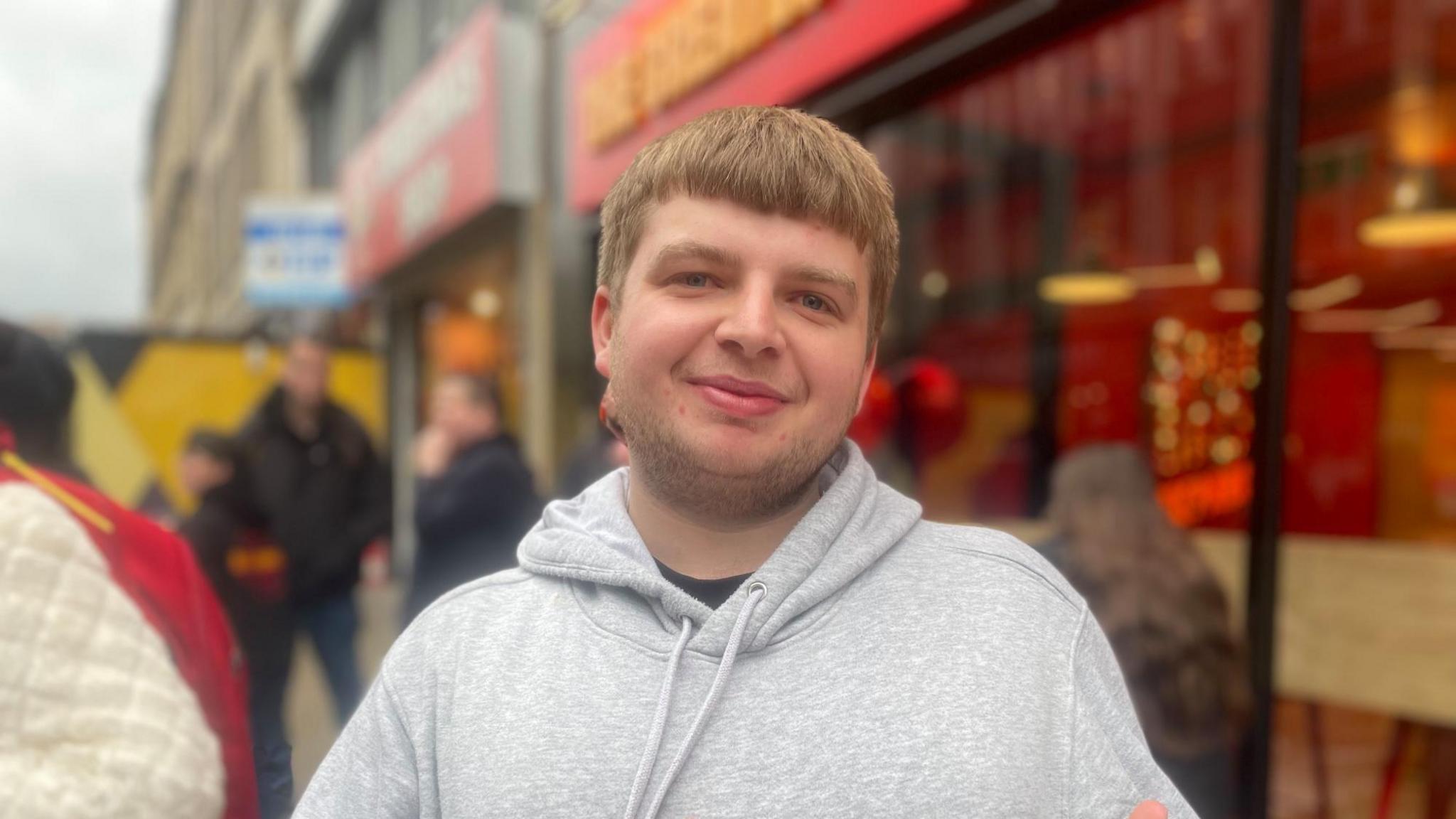 A young man with fair hair and a grey hoodie smiles at the camera