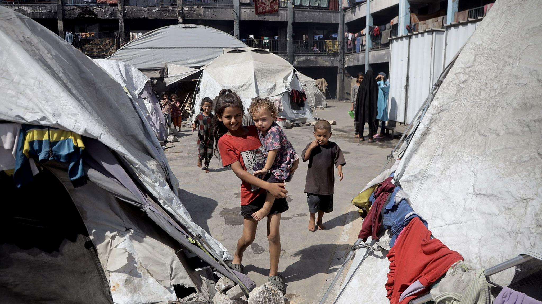 A smiling older child carries a younger girl in her arms in a displaced persons camp in Khan Younis, Gaza. The pair are surrounded by dirty grey and white tents with items of clothing drying on the outside. there are other children and women in background.