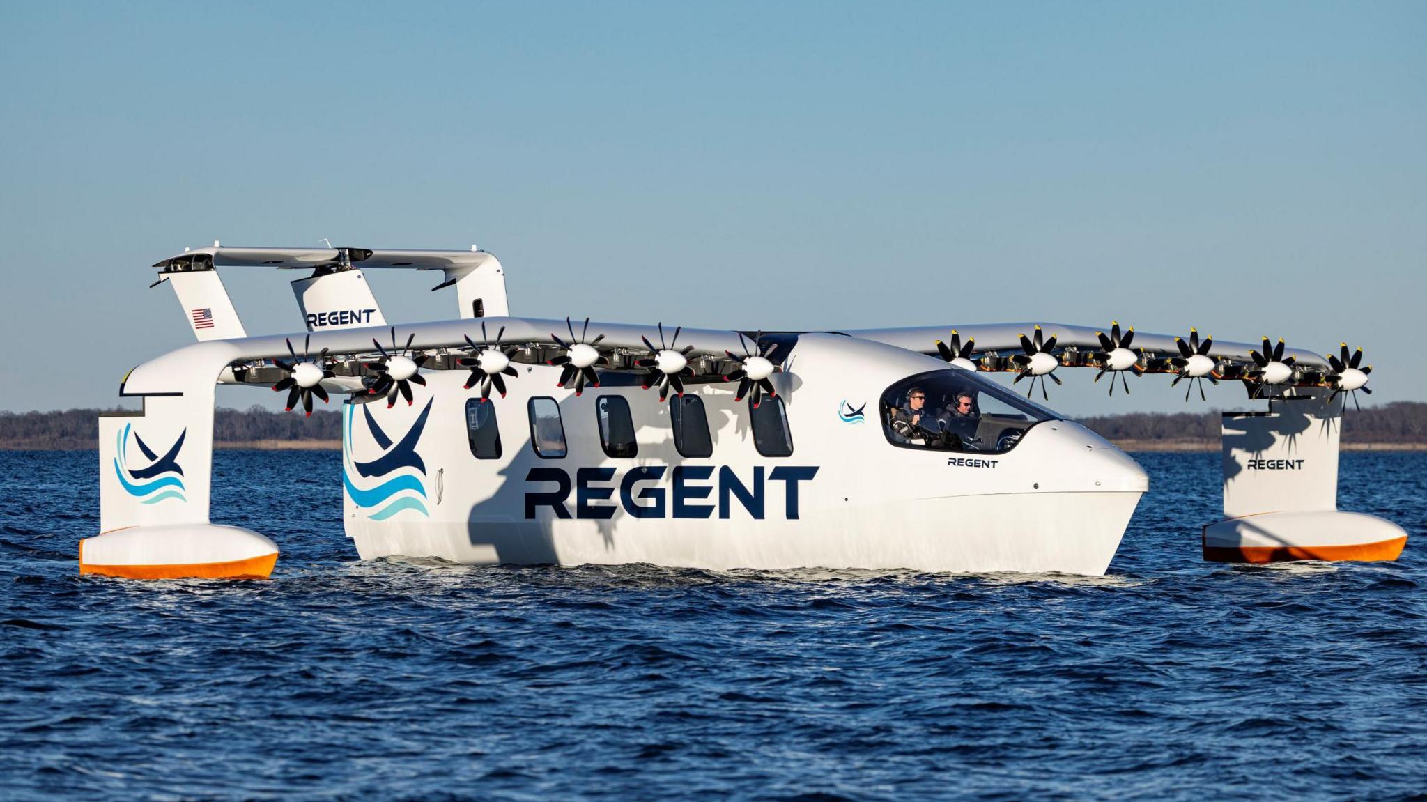 The prototype Seaglider is moving slowly across the surface of the sea on a sunny day. The machine looks like an aircraft with curved wings and six small propellers on each wing. It has hydrofoils on its undercarriage and wings. The machine is shown flying low over the surface of the sea. The fuselage is painted white and blue, and has five windows down its side and the word "Regent".