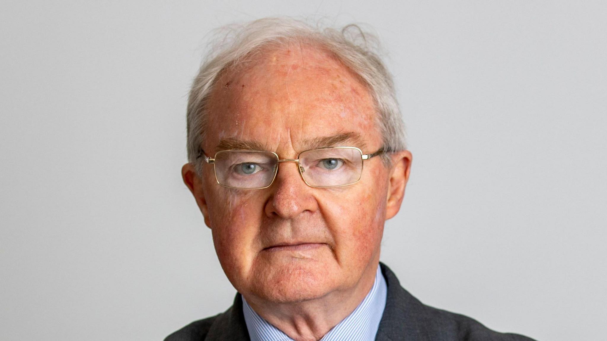 Sir Declan Morgan: a white haired man wearing gold framed glasses, looking into the camera - wearing a navy suit and striped blue shirt 