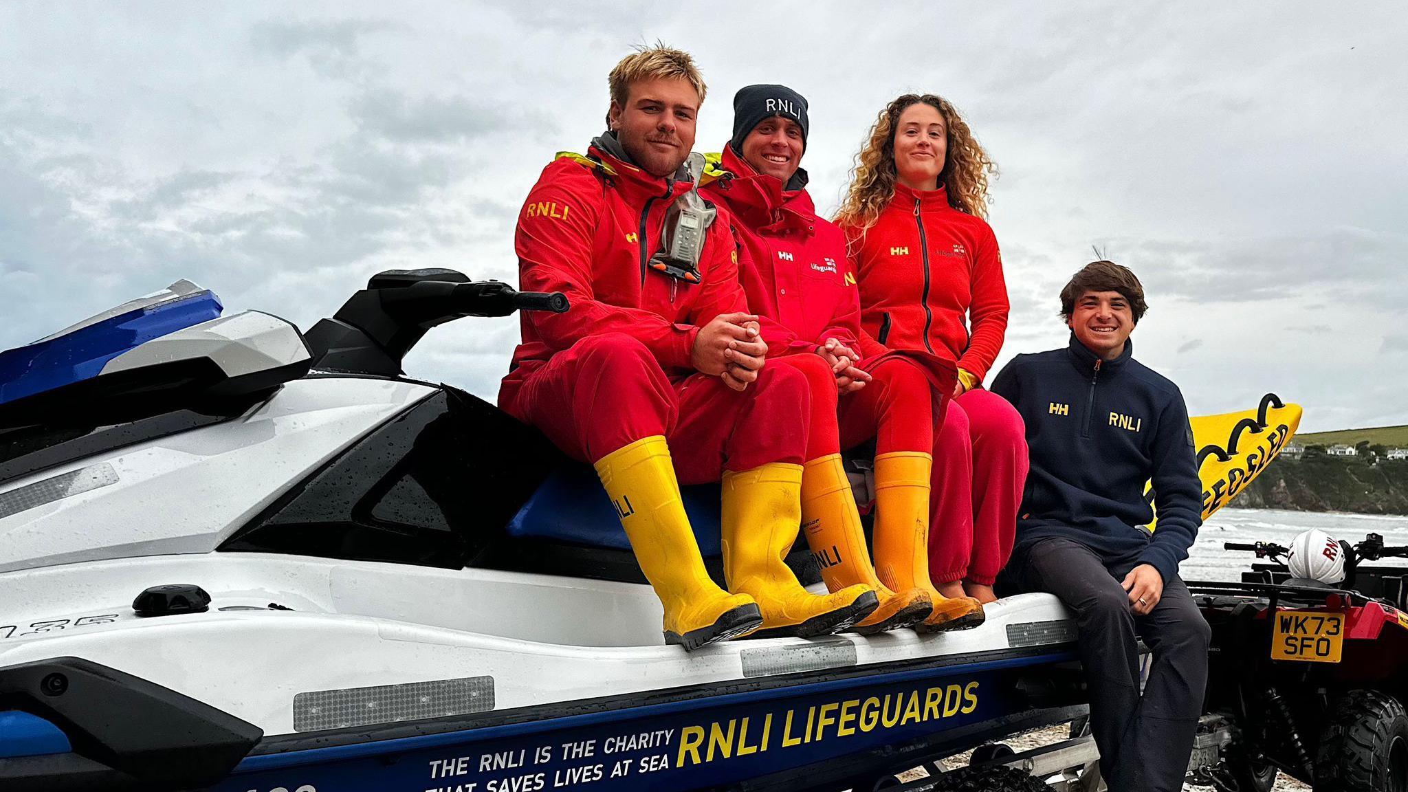 Four lifeguards involved in the rescue, sitting on a blue and white stationary rescue water craft.  from left to right is Noah Hindley, Matt Pyman, Emily Lawrence and Luke Lane-Prokopiou. The first three are wearing red jackets and trousers, with yellow wellies. The person on the right is wearing a dark jacket and trousers and is positioned lower than the first three people. 