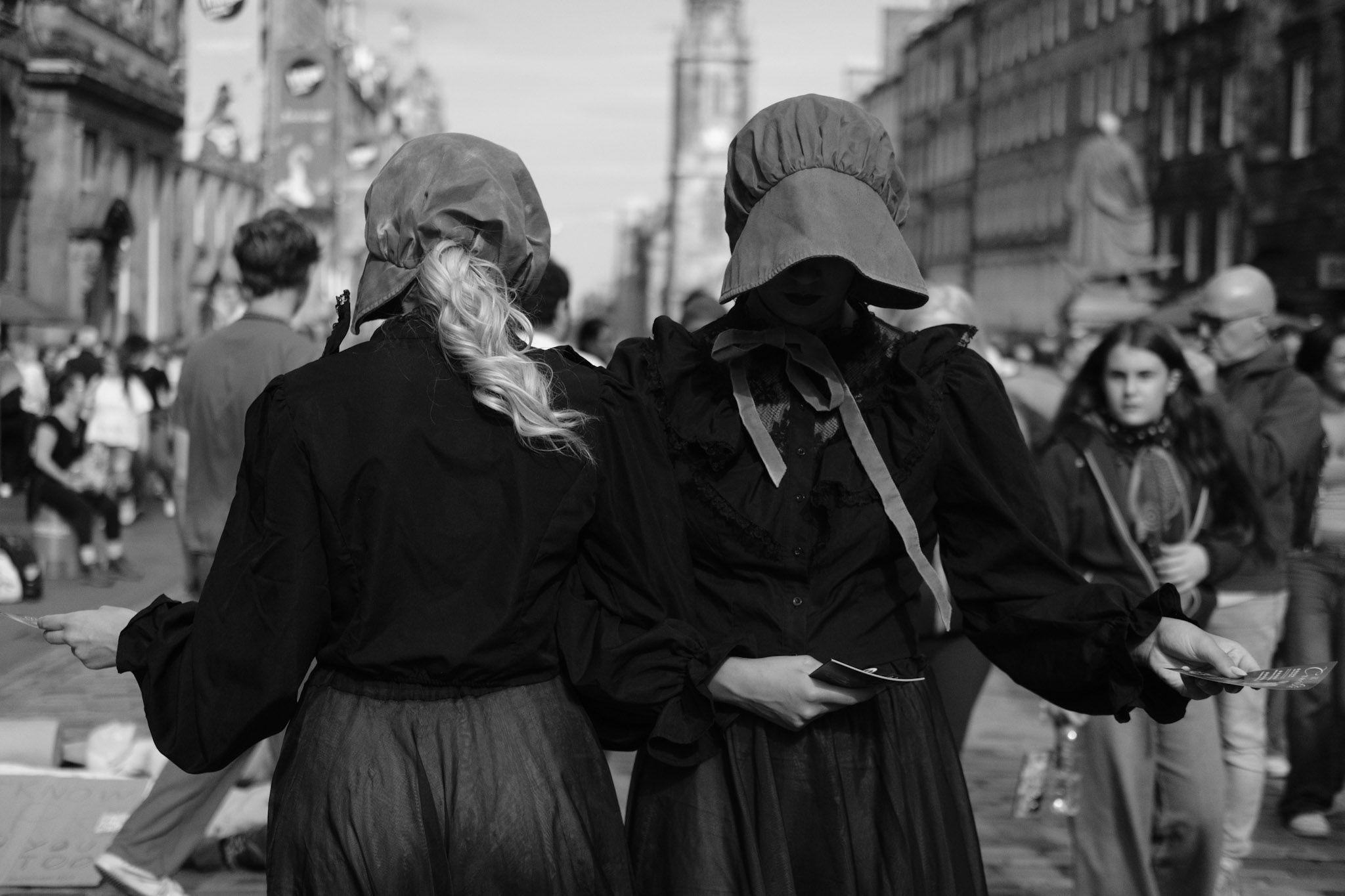 A monochrome image of two women, facing opposite directions and wearing bonnets with their faces covered, hand out promotional materials in the street.