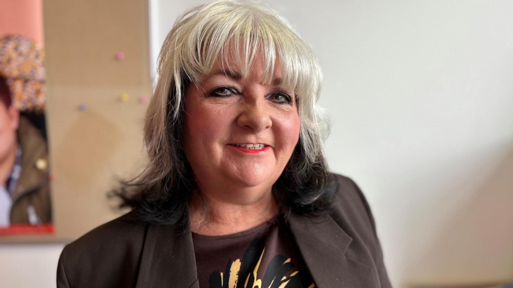 Head-and-shoulders portrait of a person with shoulder-length two-tone hair, wearing a dark blazer over a patterned top. The person is facing the camera indoors, with a noticeboard and softly blurred background visible behind them.
