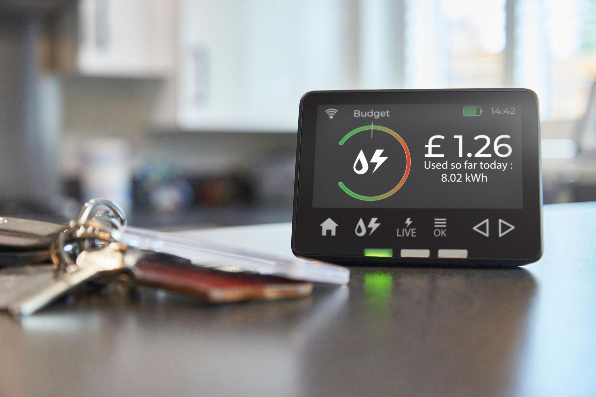 A modern smart meter displaying the number of units used sits on a kitchen counter. A set of keys is on the counter beside it.