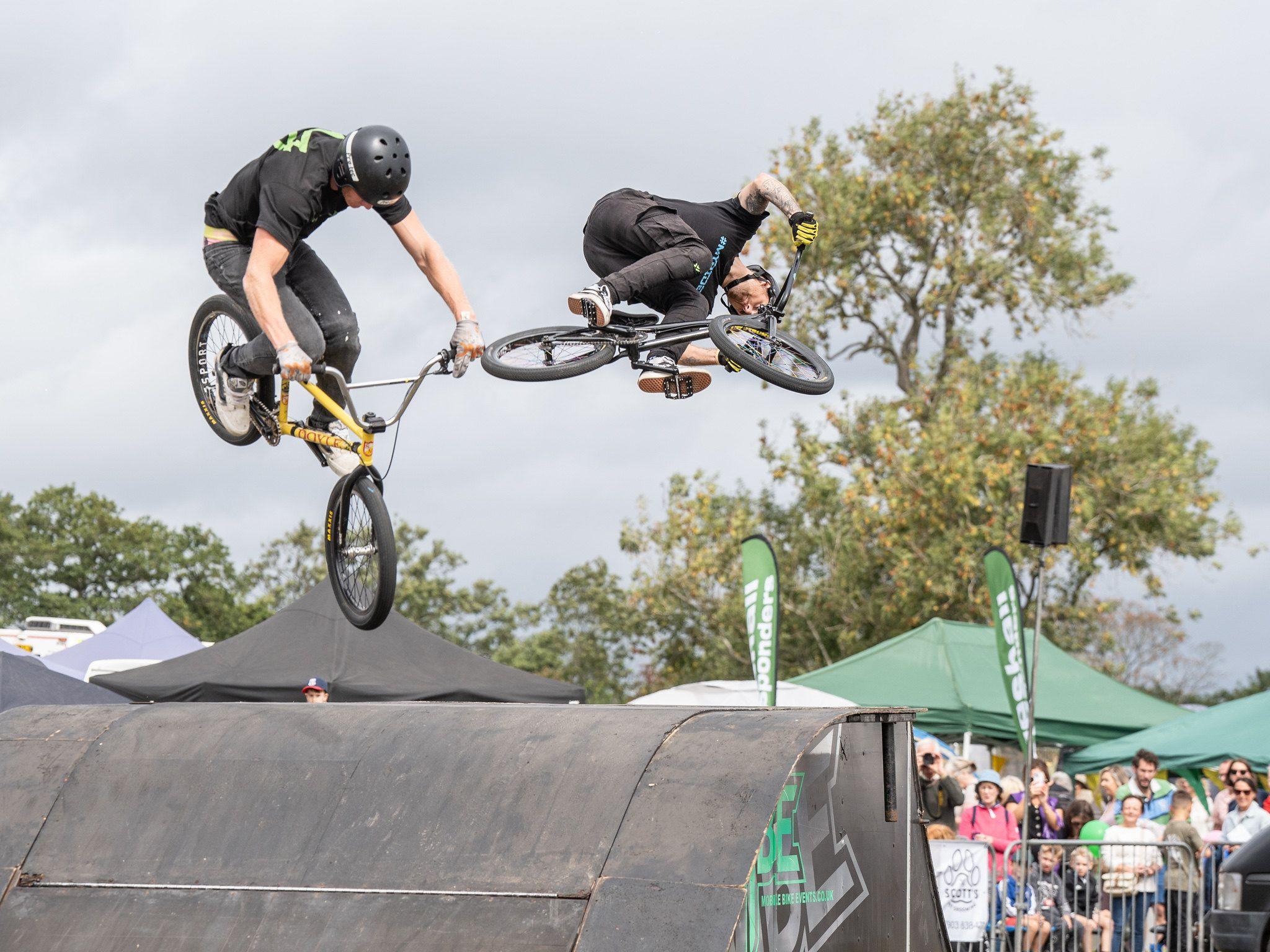 Two BMX riders are seen in the air above a ramp as an audience looks on. They are both wearing helmets and dark tee-shirts.