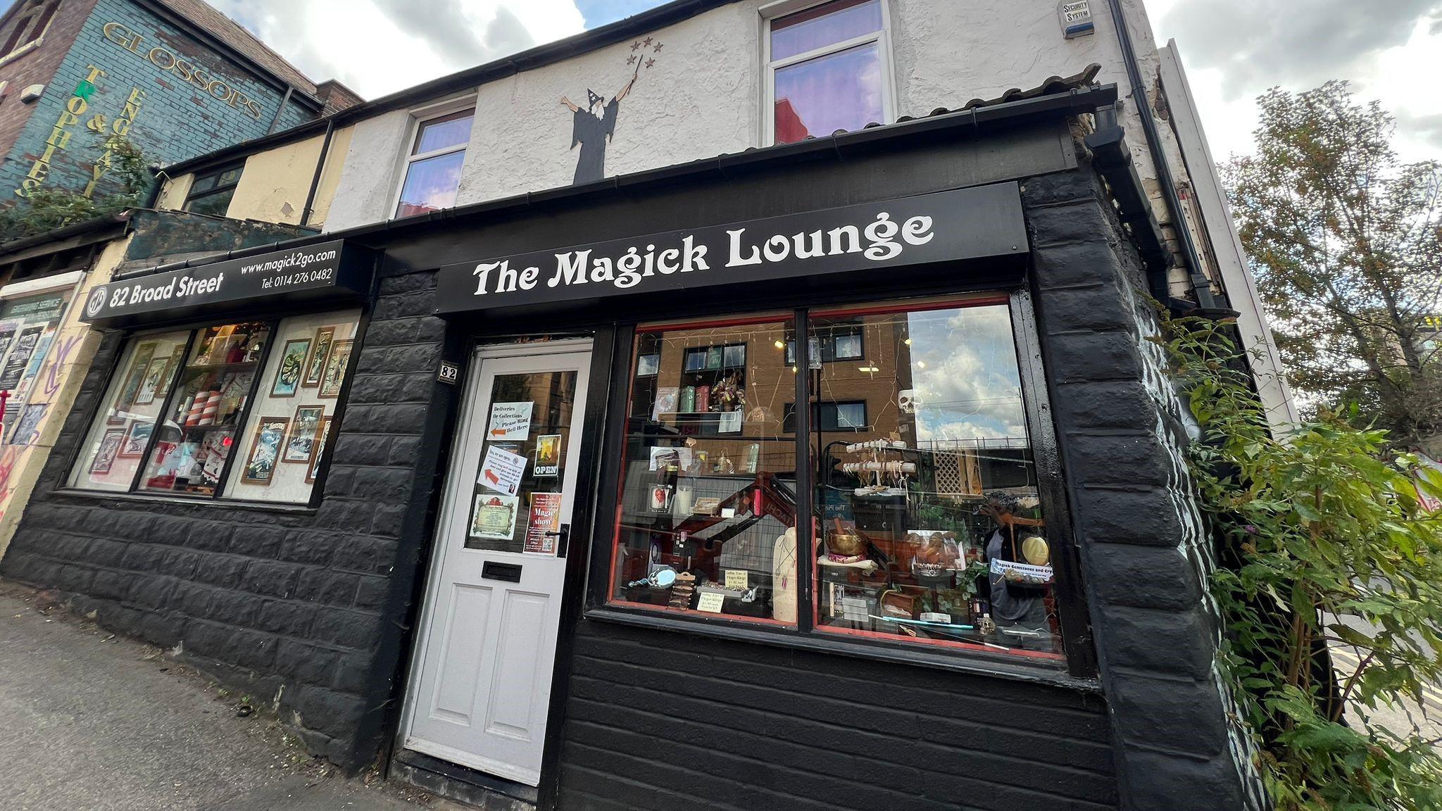 A street-level view of a small shop called The Magick Lounge. The building has a black-painted lower facade with a white door and large front window displaying various items. Above the shop sign, the upper part of the building is light-coloured with two windows and a mural of a silhouetted magician holding a wand.