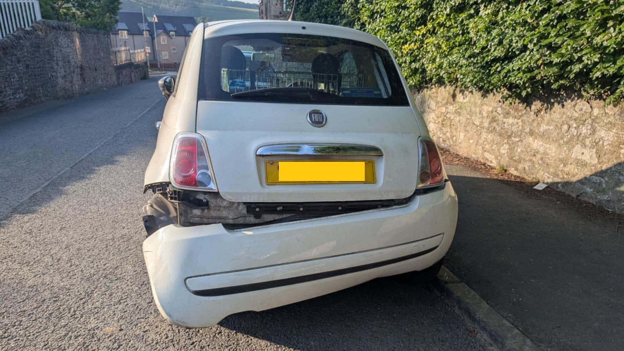 A white Fiat car, parked next to a pavement, with its rear bumper hanging off