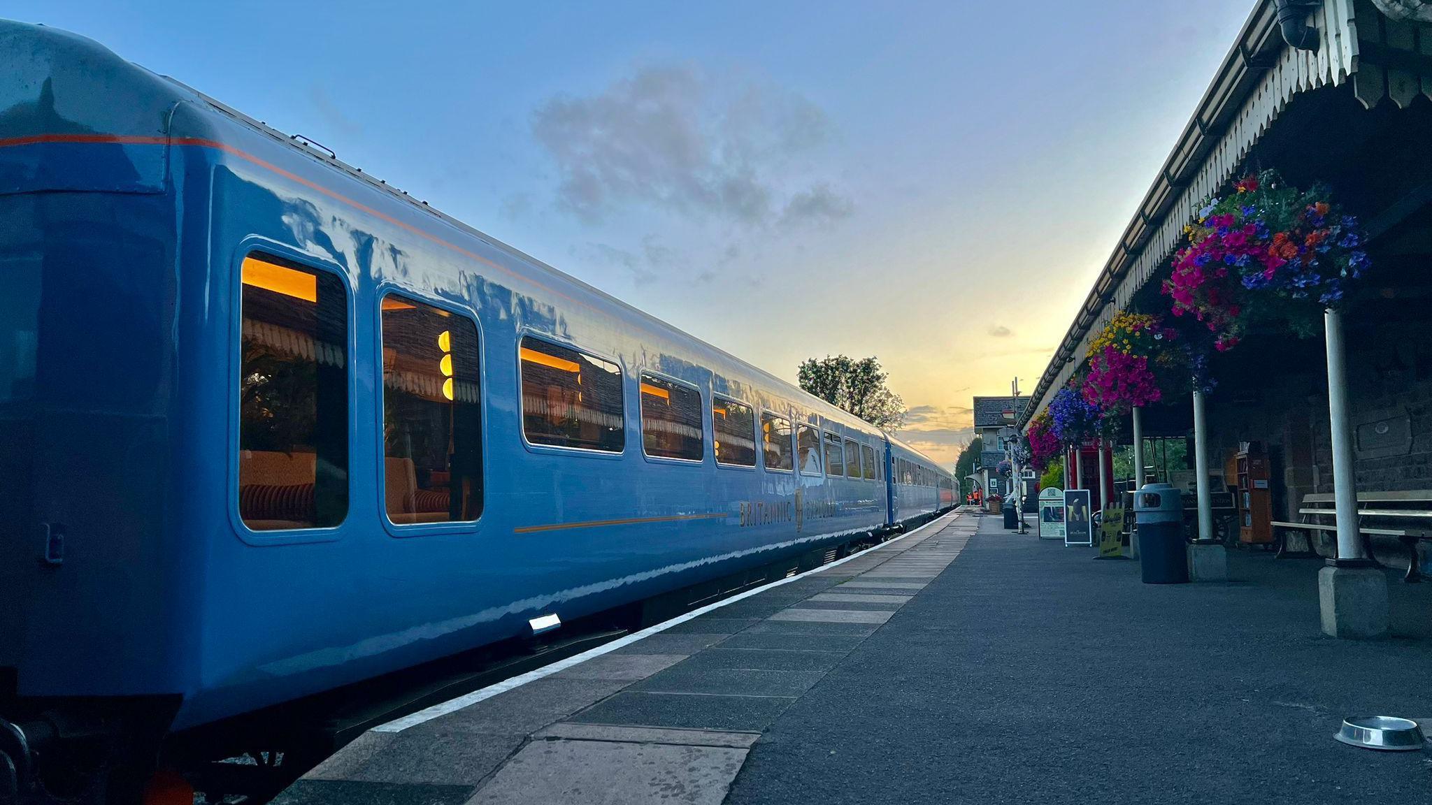 A vintage blue train carriage is parked by a platform as the sun sets.