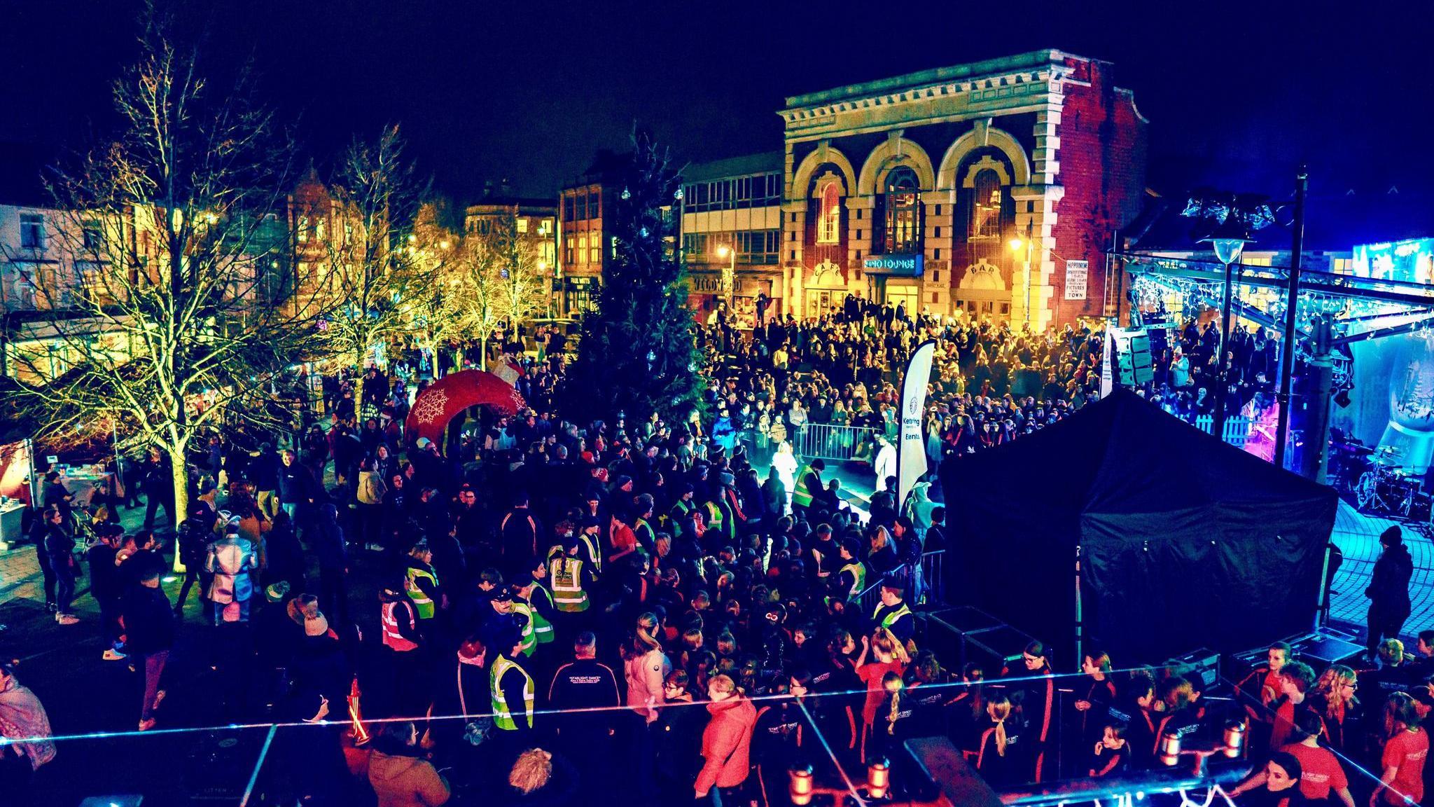 A photo of Kettering town centre with buildings illuminated in lots of different coloured lights. Large crowds are gathered around a large Christmas tree.