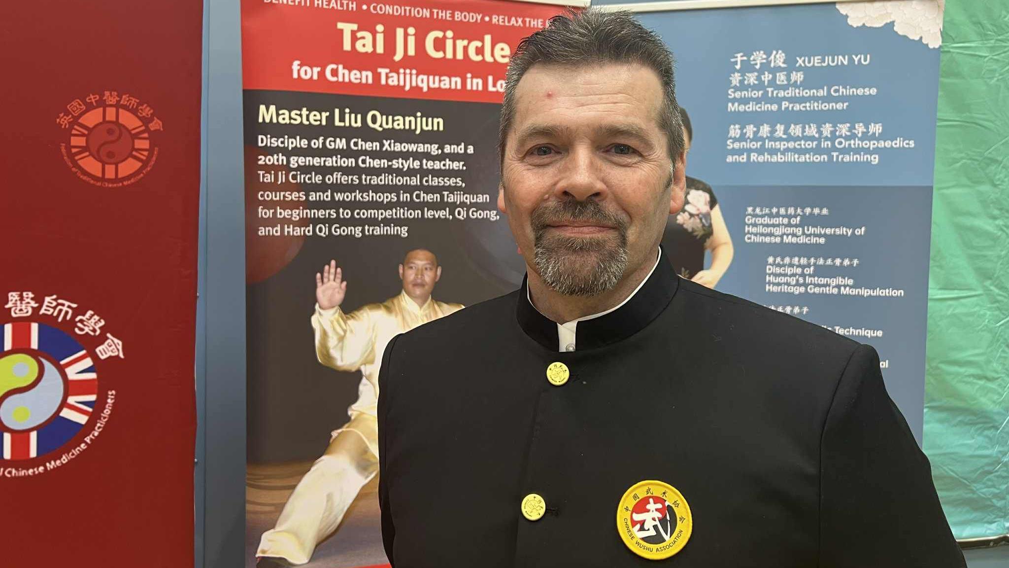 A man in a black, traditional martial arts uniform with yellow buttons and a badge stands in front of informational banners at an event.
