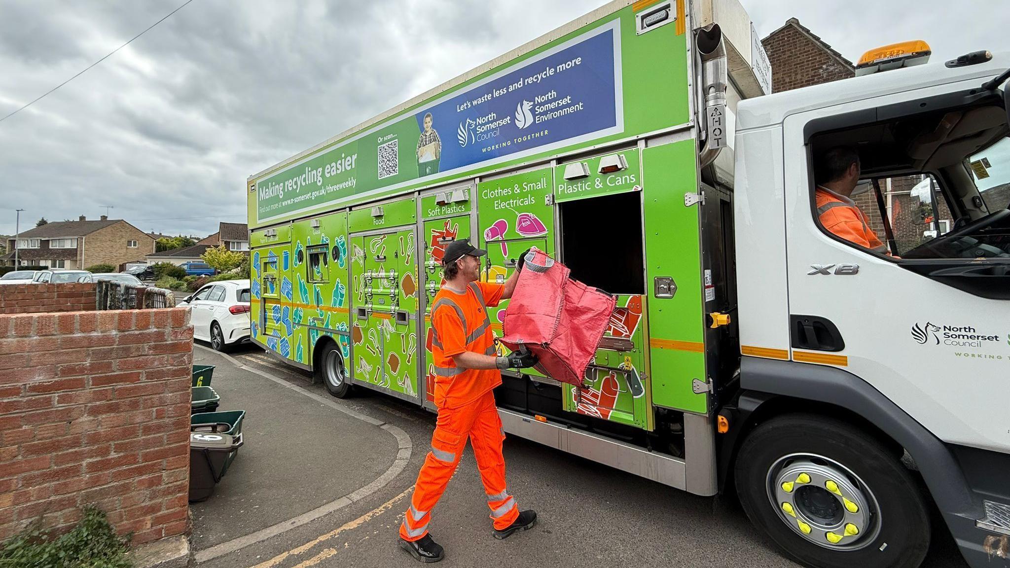 A refuse worker empties one of the new large red plastic recycling bags into a green recycling truck parked on a residential street.