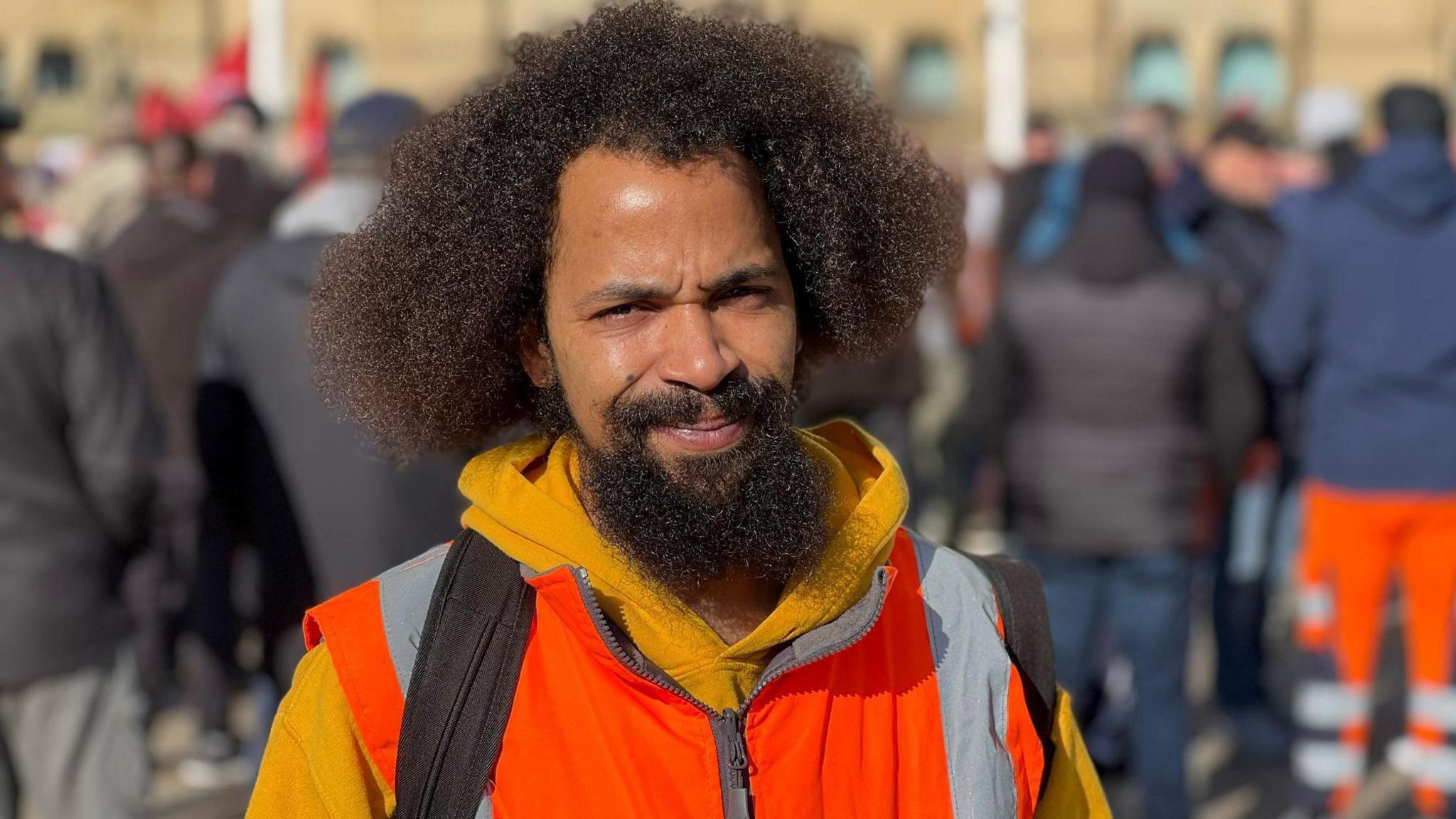 Steevn is looking at the camera and had a light orange jumper and is wearing an orange high-vis jacket. He has Afro hair and a beard and a bag on his back. Behind him the backs of protesters can be seen blurred out. 