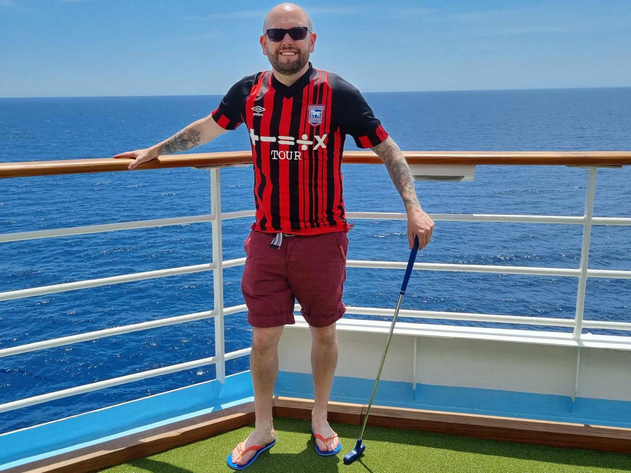 Chris Peachey on a cruise ship, wearing a red and black Ipswich Town away kit