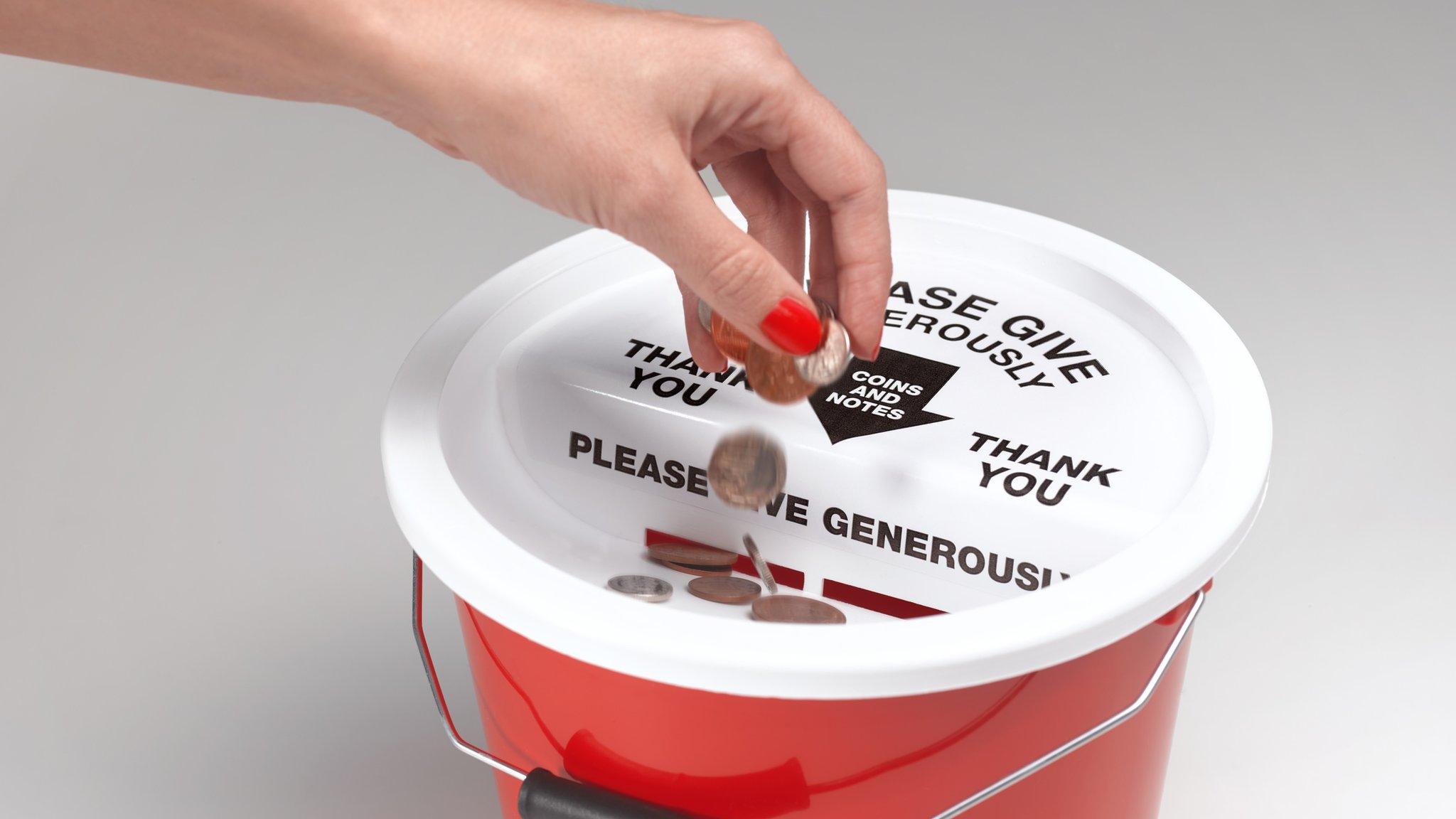 A hand drops coins into a red charity bucket