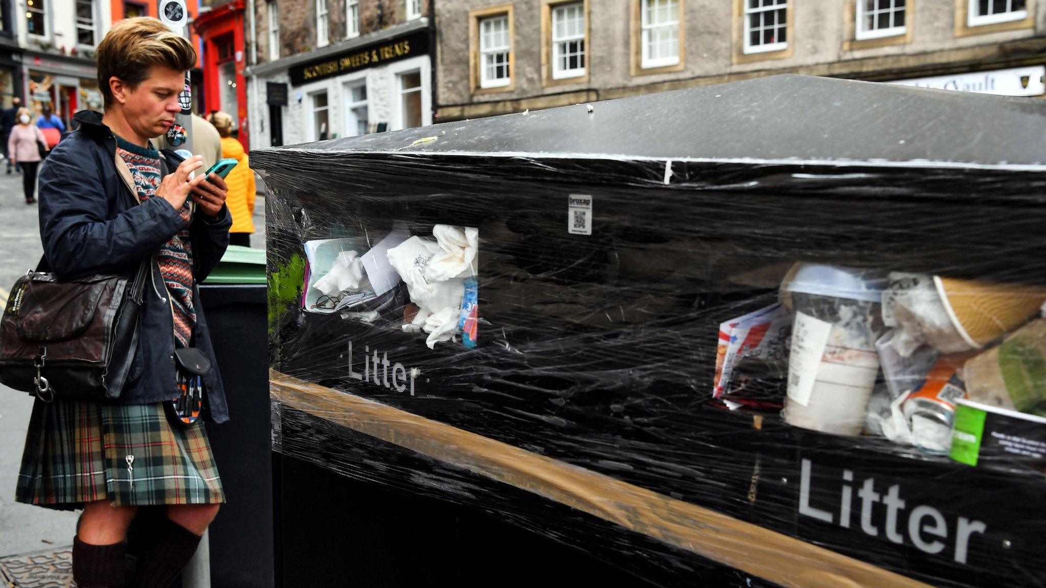 a person stands near full bins in edinburgh which have been wrapped in cling film to stop people throwing their litter away