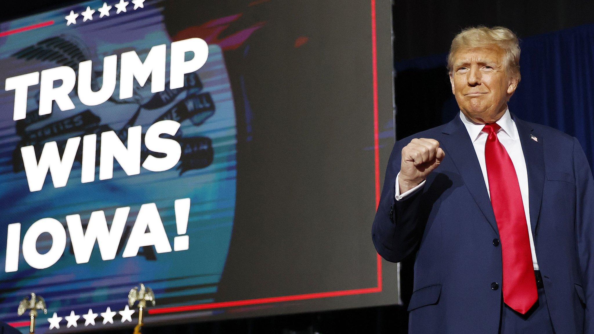 trump holds his fist up while smiling on stage at the iowa caucuses with a sign behind him which reads trump wins iowa