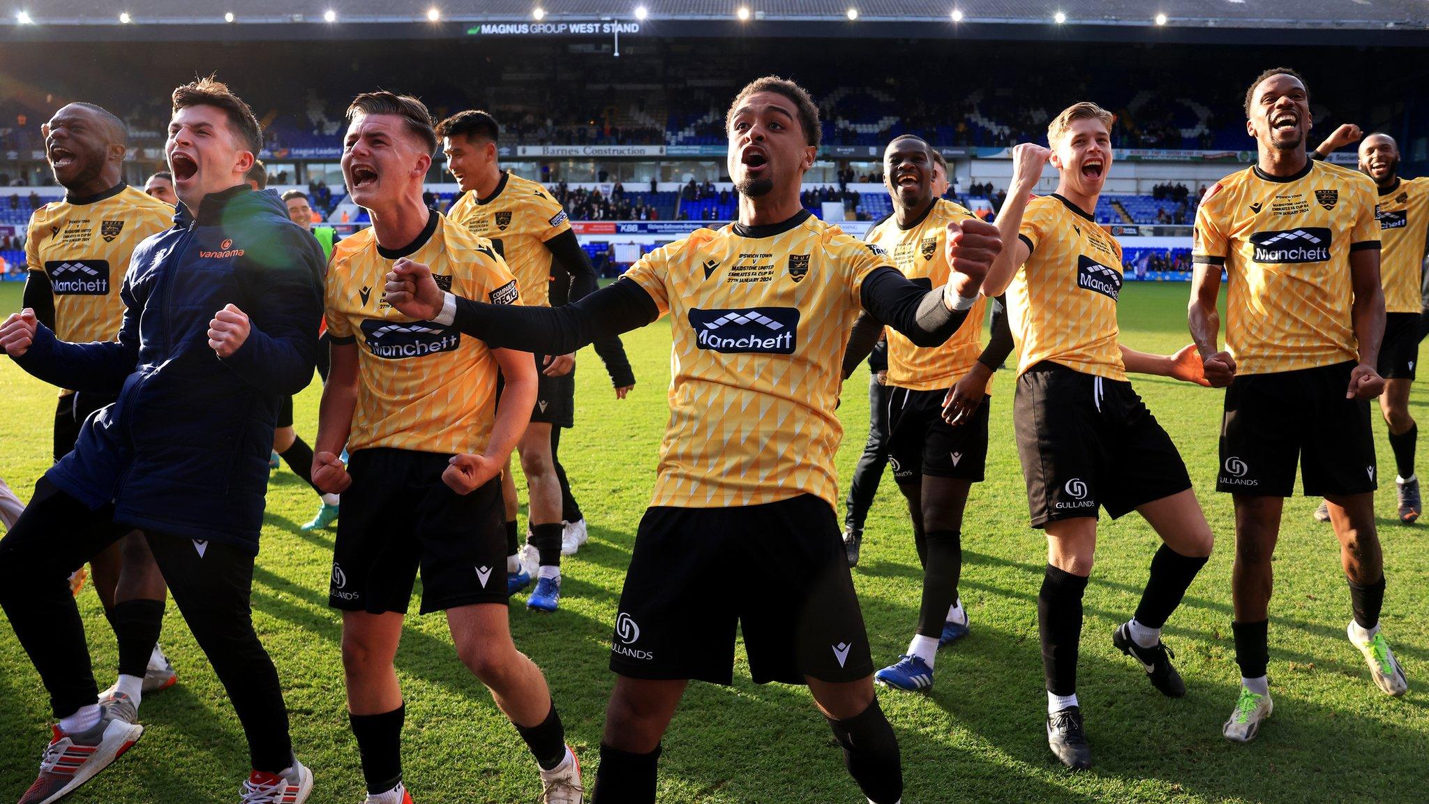 Maidstone United players dressed in black and yellow kits celebrate in a large group on the pitch in front of the camera