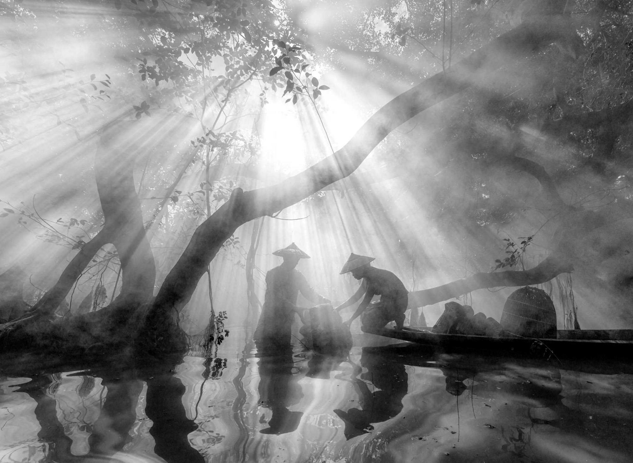 A black an white image showing two local fishermen surrounded by trees and water. The light is streaming through the branches and the fishermen are reflected in the water.