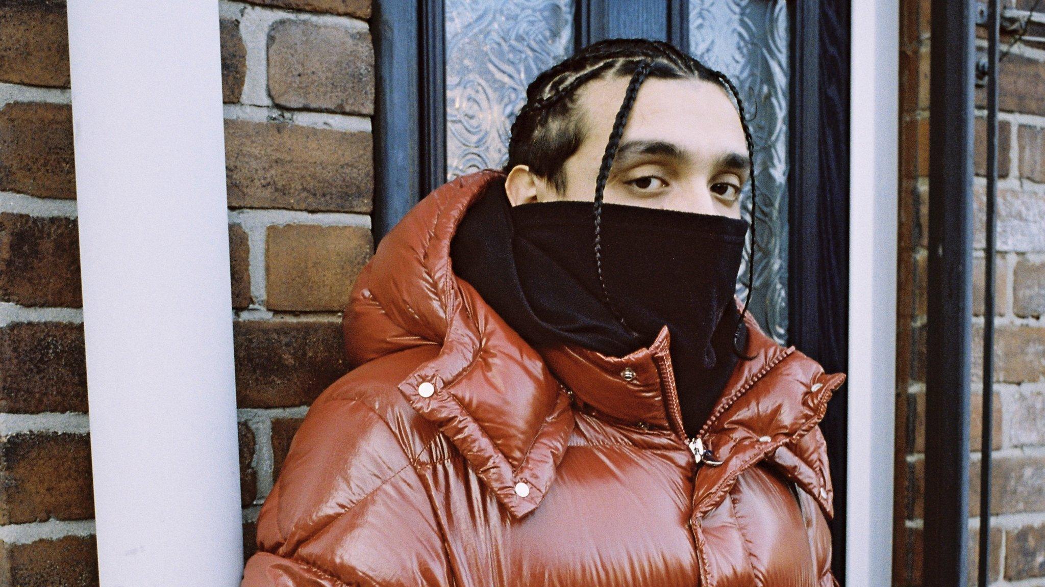 Sliime standing against a wall. He's wearing a red coat and his face is partly covered, his hair is in braids