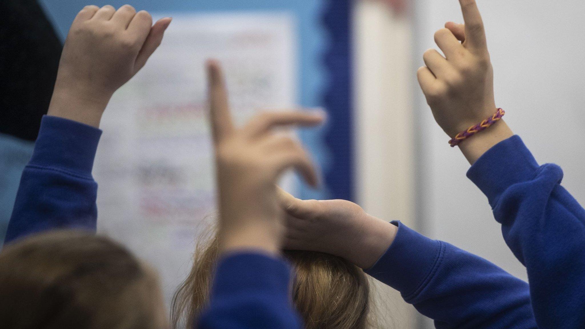 Young children with their hands up in a classroom. They they wearing dark blue jumpers and white shirts