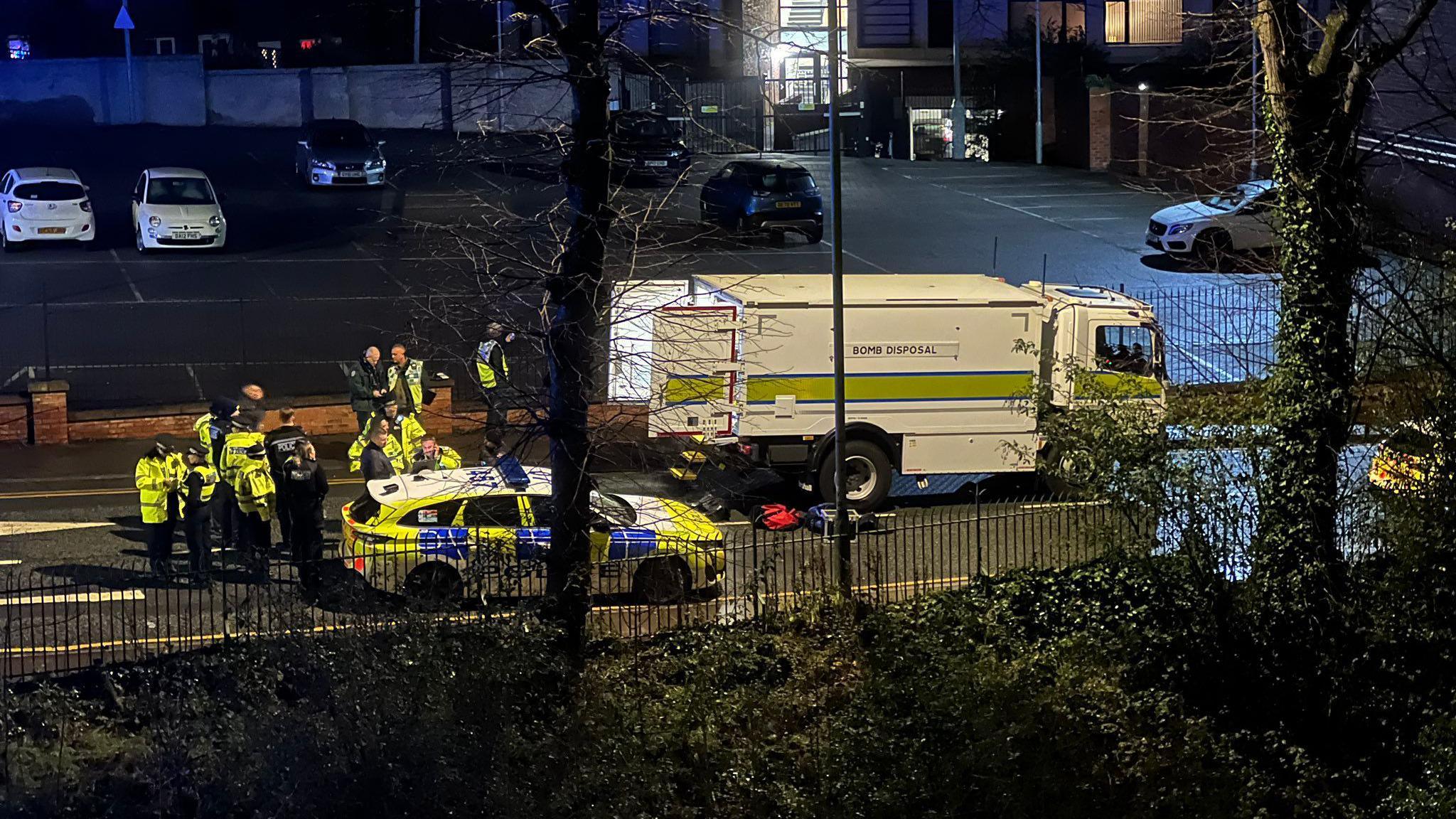 A bomb disposal van with the rear doors open next to a police vehicle with a group of police in high vis seen through the night sky. 