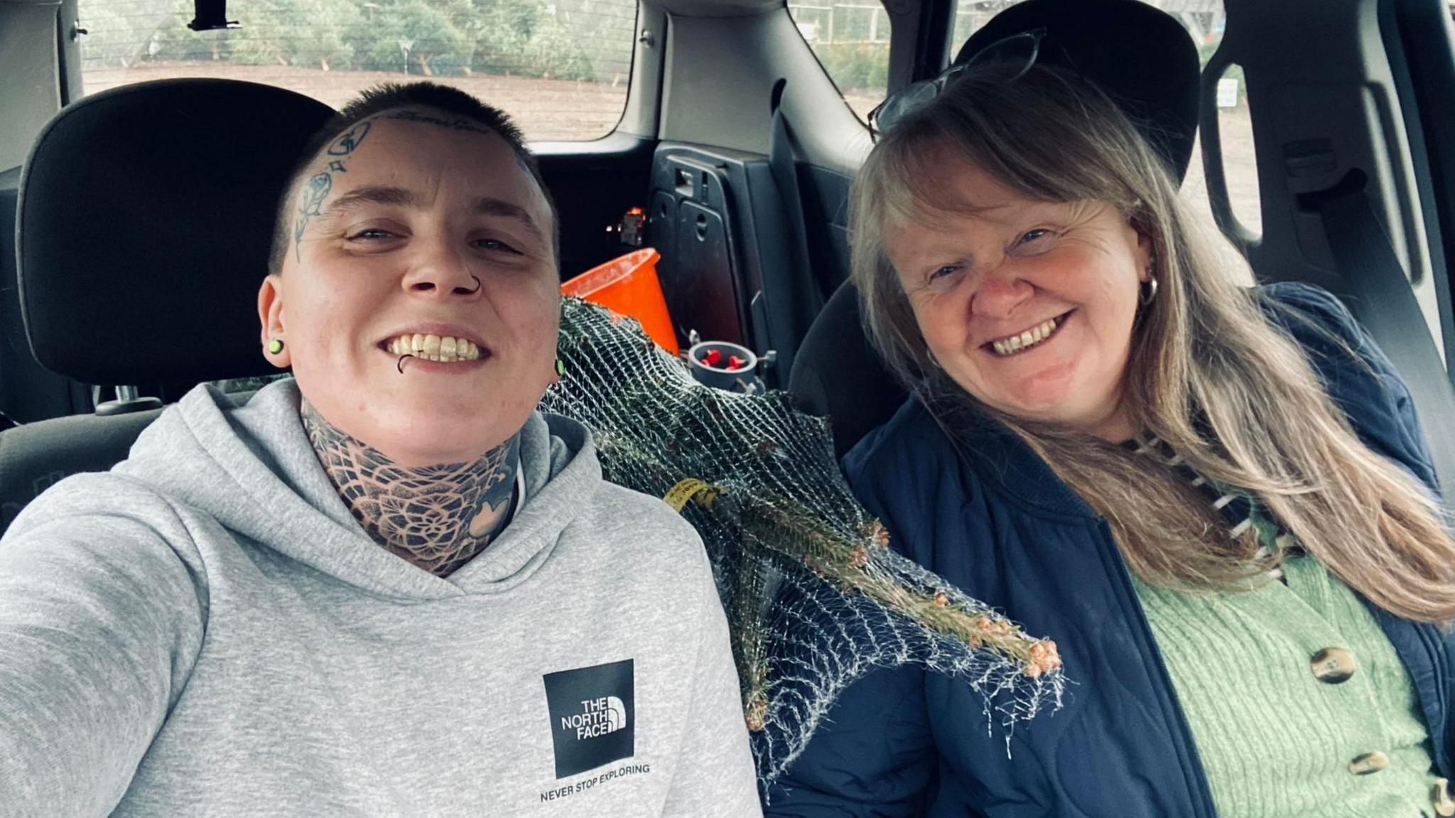 Noah and Sara taking a selfie in a car, with a Christmas tree between them. Noah has facial and neck tattoos, and a lip piercing, and is wearing a grey hoodie. Sara is on the right, has a green and blue top on and long brown hair.