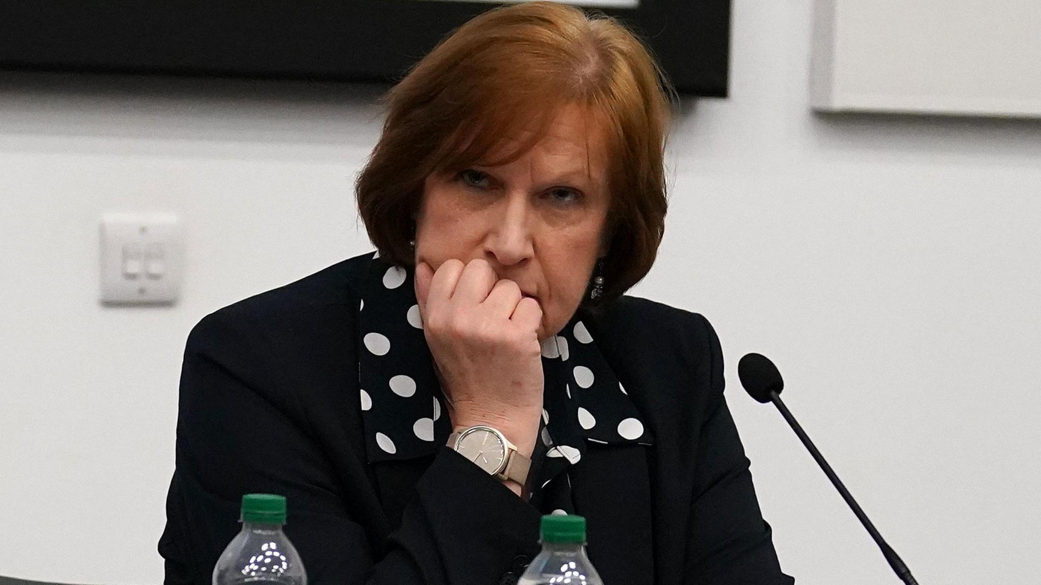 A woman, wearing a black blazer and black and white polka dot blouse, seated at a table. She is resting her chin on her hand. In front of her is a microphone, two water bottles.