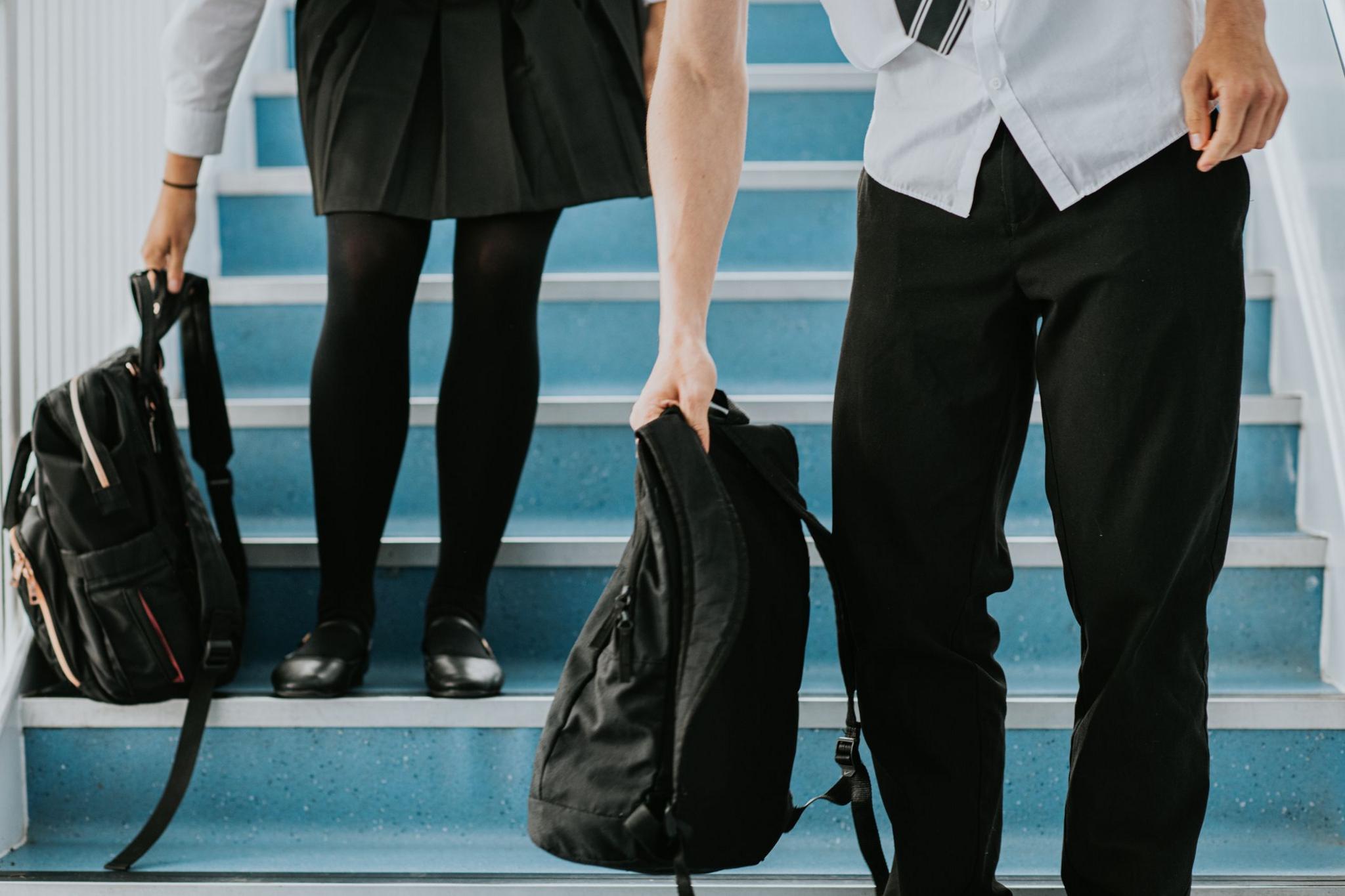 A girl wearing a school uniform skirt and a boy in trousers stand on steps inside a school corridor.