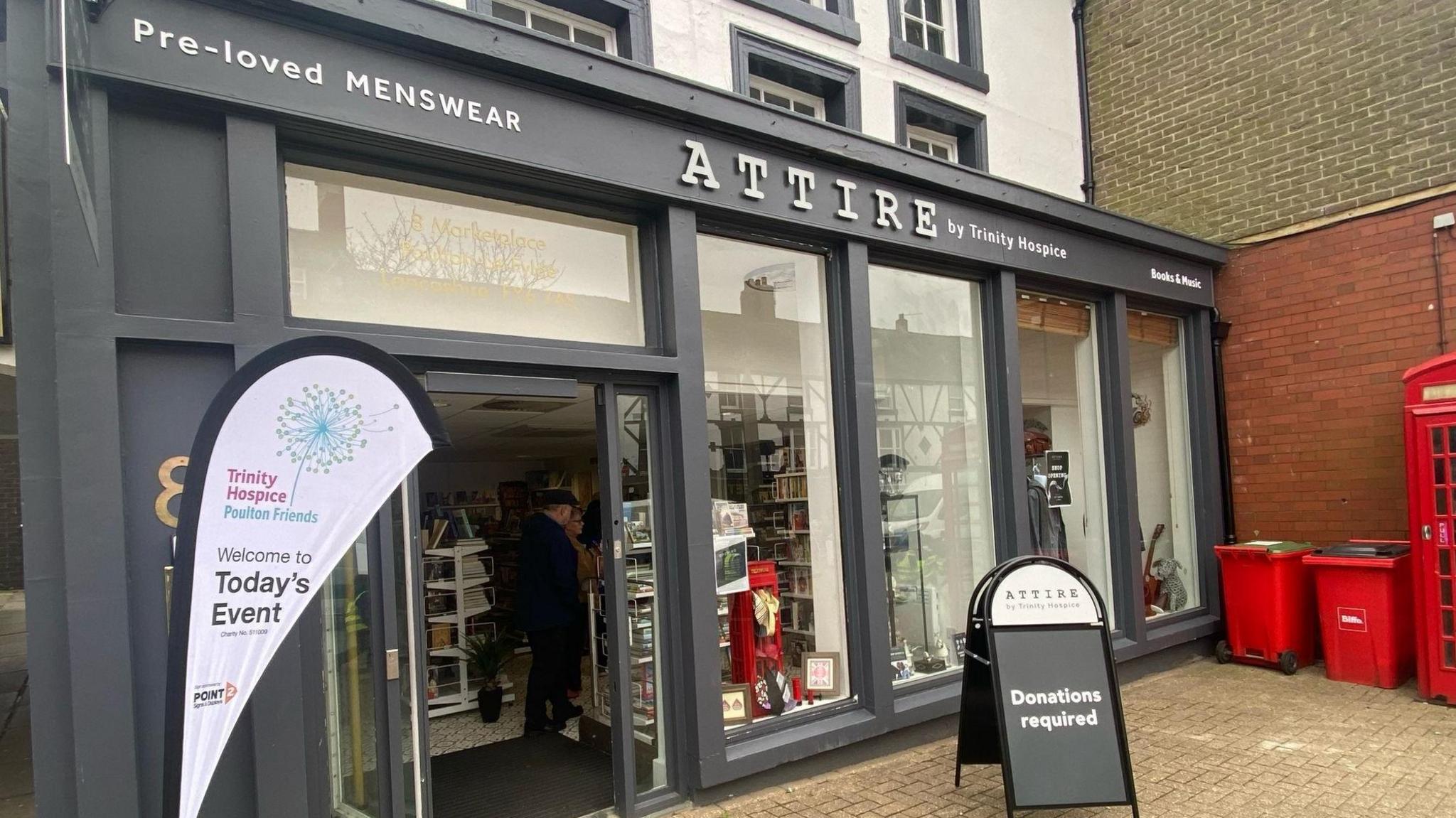 A glass-fronted shop with contemporary grey window and door frames. A sign reading 'donations required' can be seen outside while customers can be seen milling around in the entranceway. 