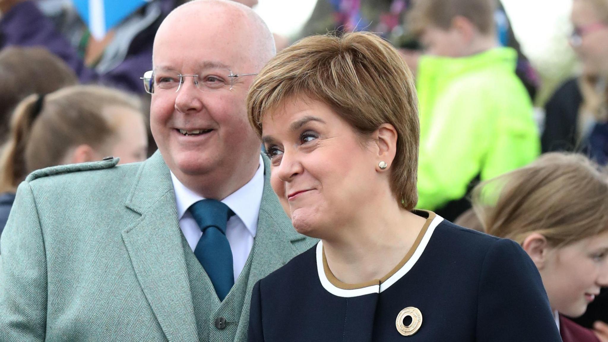 Peter Murrell and Nicola Sturgeon before at an event their separation. Murrell has a bald head and is wearing thin-framed glasses, a green jacket and waistcoat with a white shirt and blue tie. Sturgeon is smiling and is wearing a blue dress. There are children in the background