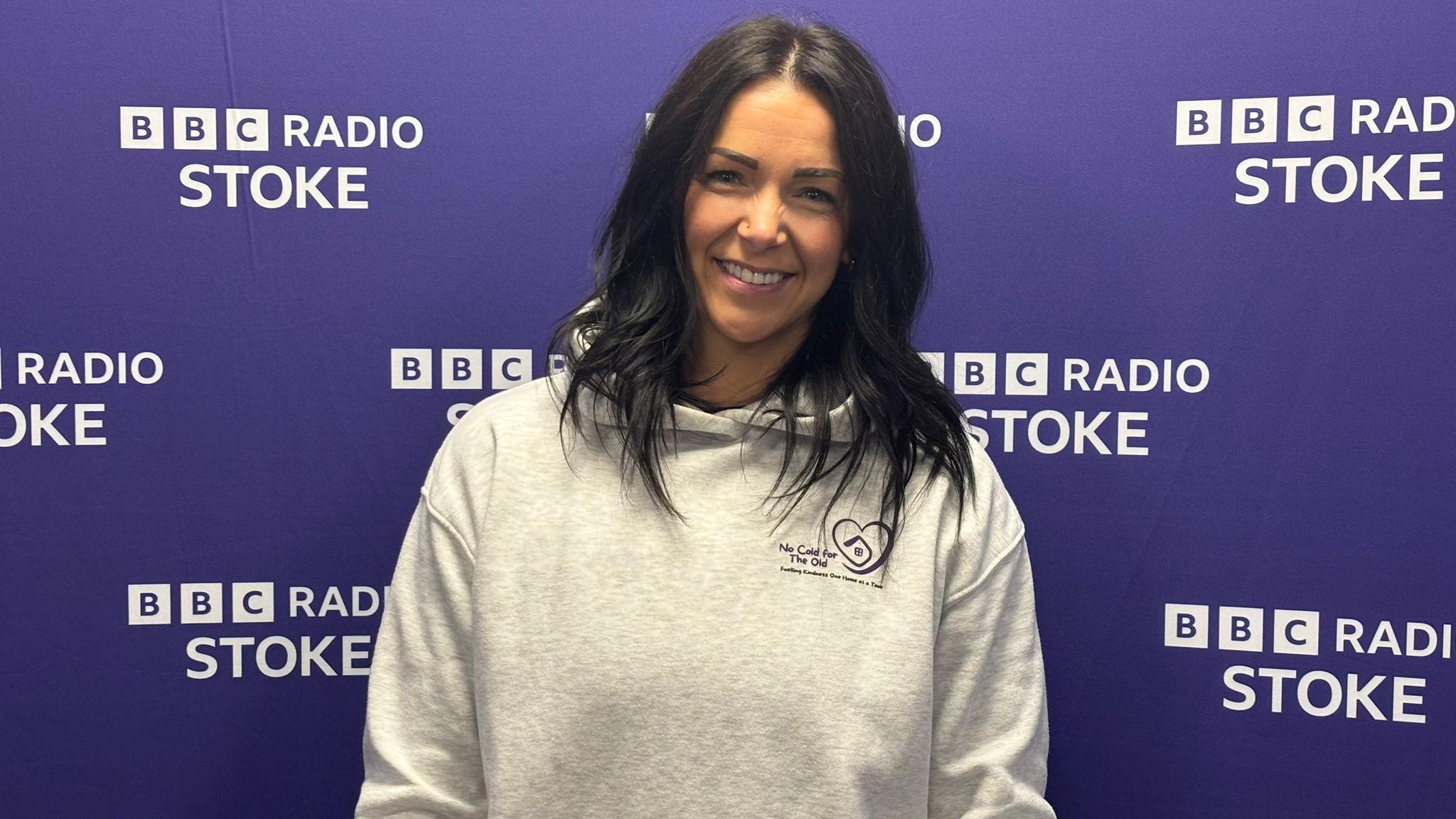 A woman with long black hair and a light grey hoodie smiles as she stands in front of a purple background with the BBC Radio Stoke logo on it.