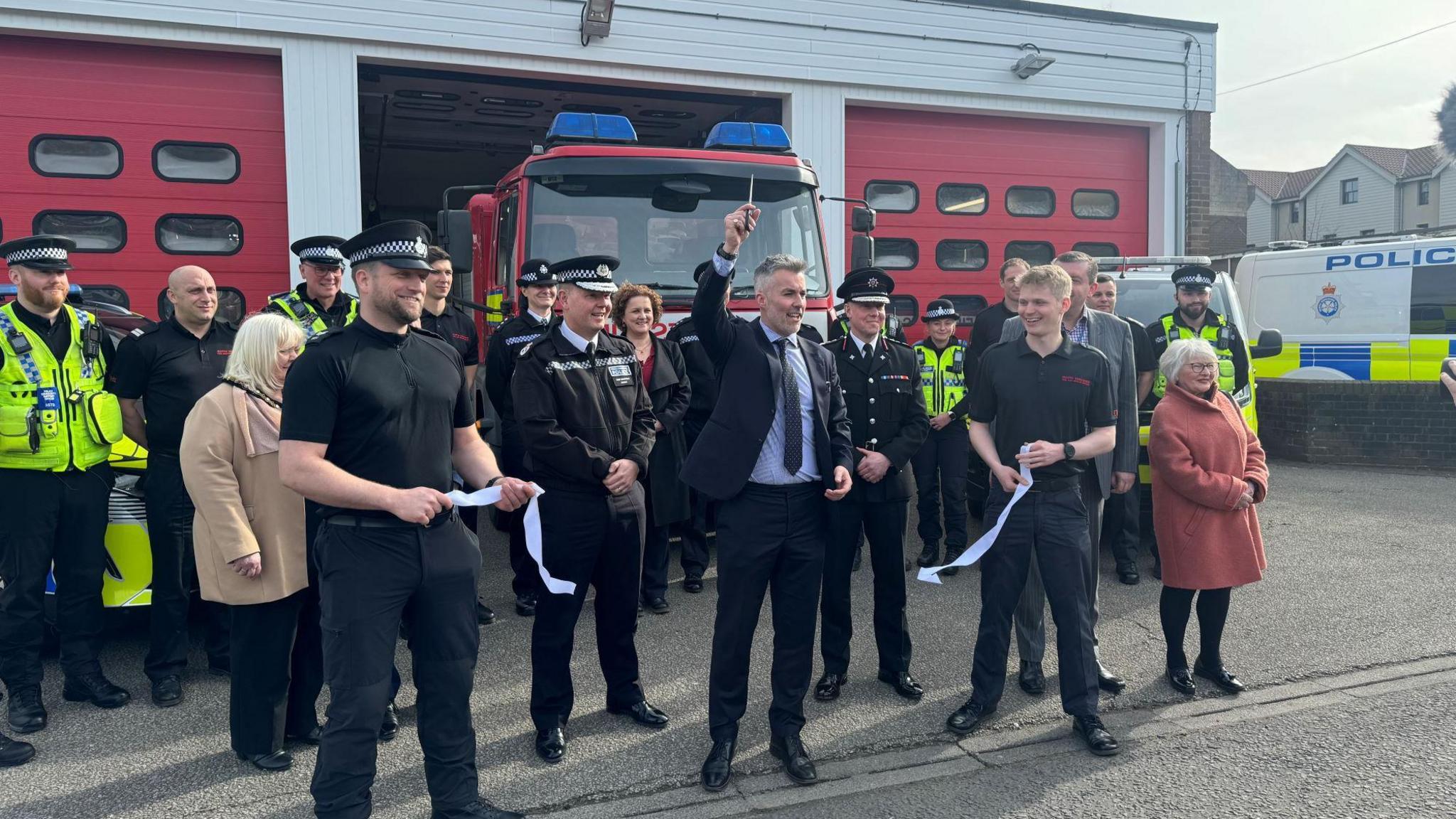 Police officers and firefighters stand outside a station. Mayor David Skaith holds a pair of scissors in the air, after cutting a white ribbon.