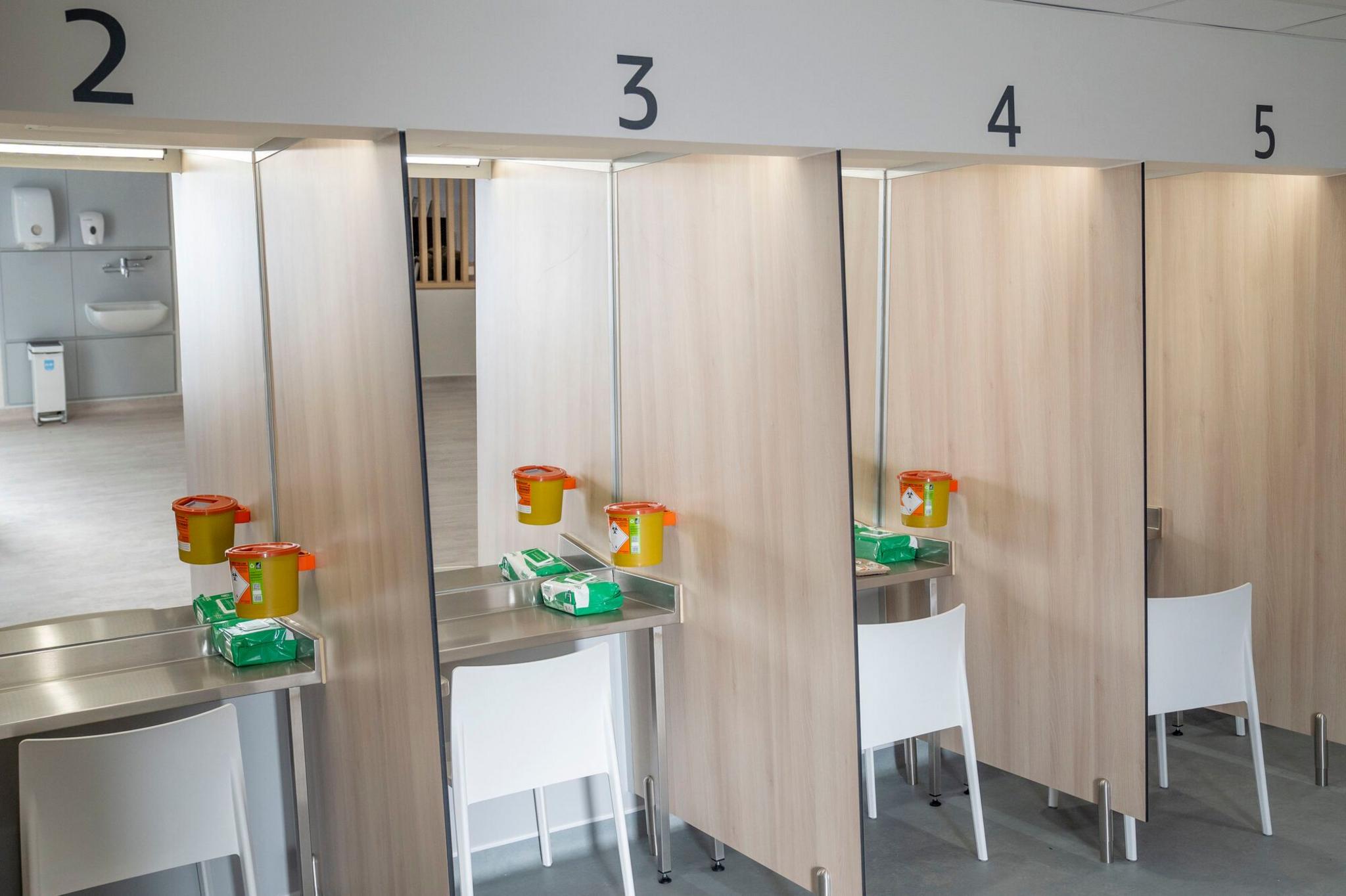 Numbered booths with a white chair in each booth. Each one faces a mirrored wall. Each booth has a green pack and a yellow sharps bin. It is brightly lit.
