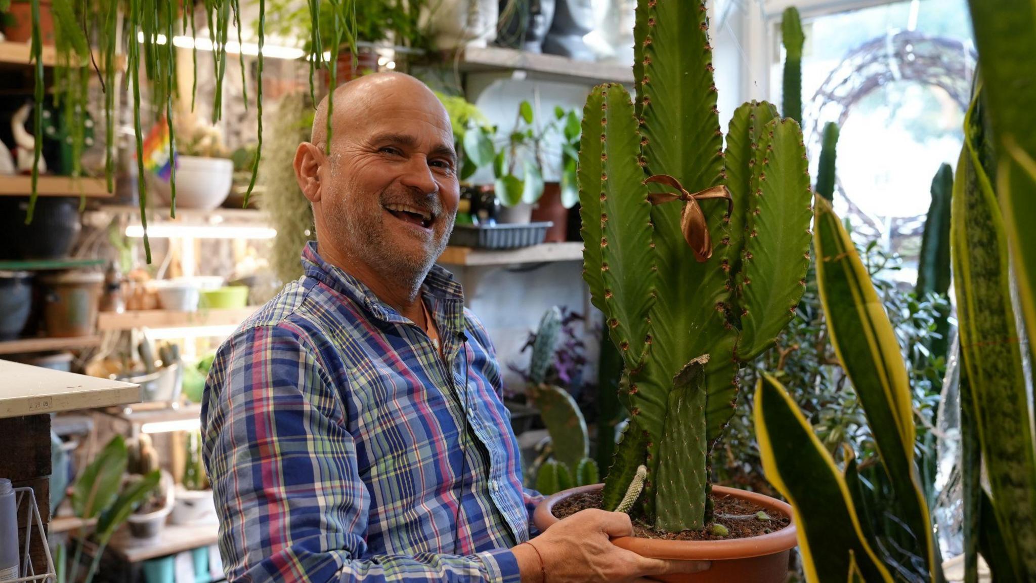 Pepe, owner of plant store Pepe Cactus in Corp Market, Canton. He holds a cactus in his hands and wears a blue and pink check shirt. Pepe has a grey short trimmed beard, a bald head and smiles boldly at the camera. Hanging plants hover above his head, while plants and cactus can be seen on lit shelves in the background. 