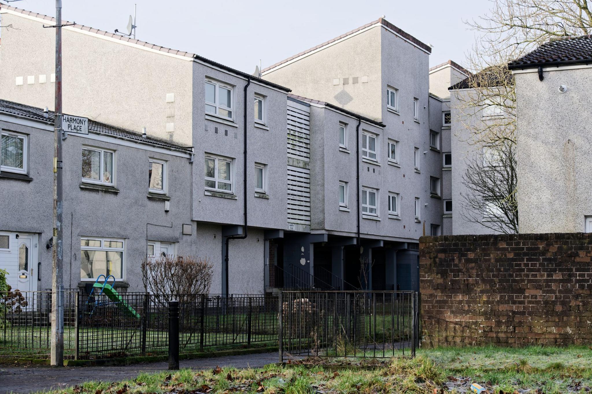 A shot of a row of local authority terraced houses and flats in Glasgow's Maryhill. A small patch of grass sits in front of the grey buildings and white wooden panels on the flats look weather-worn.