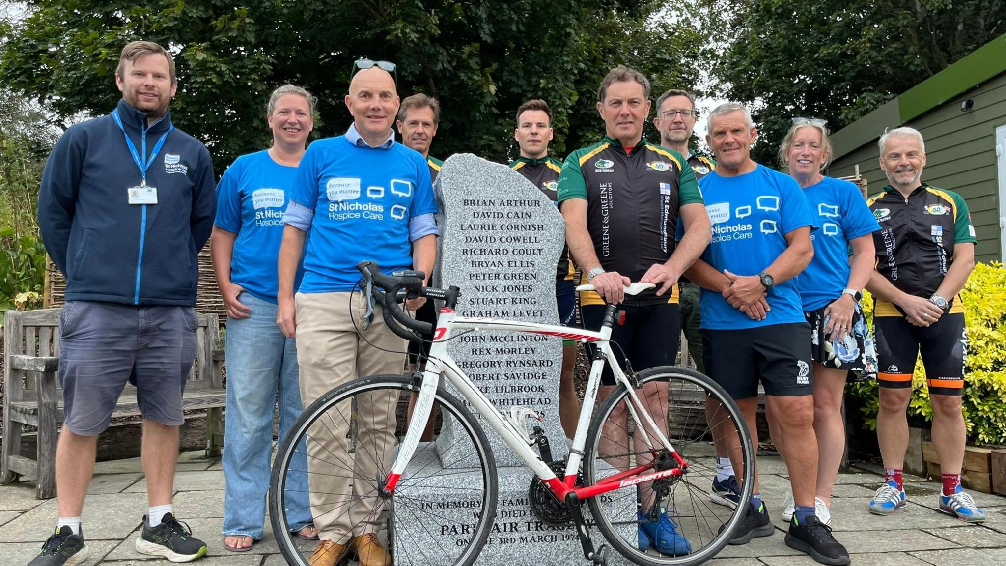 Cyclists stand in front of a stone memorial at Bury St Edmunds rugby club. Many are wearing blue t-shirts for St Nicholas Hospice - a charity benefitting from the ride. Others are in black and green cycling shorts and t-shirts. The stone has the names of victims etched into it and a bike is being held in front. 