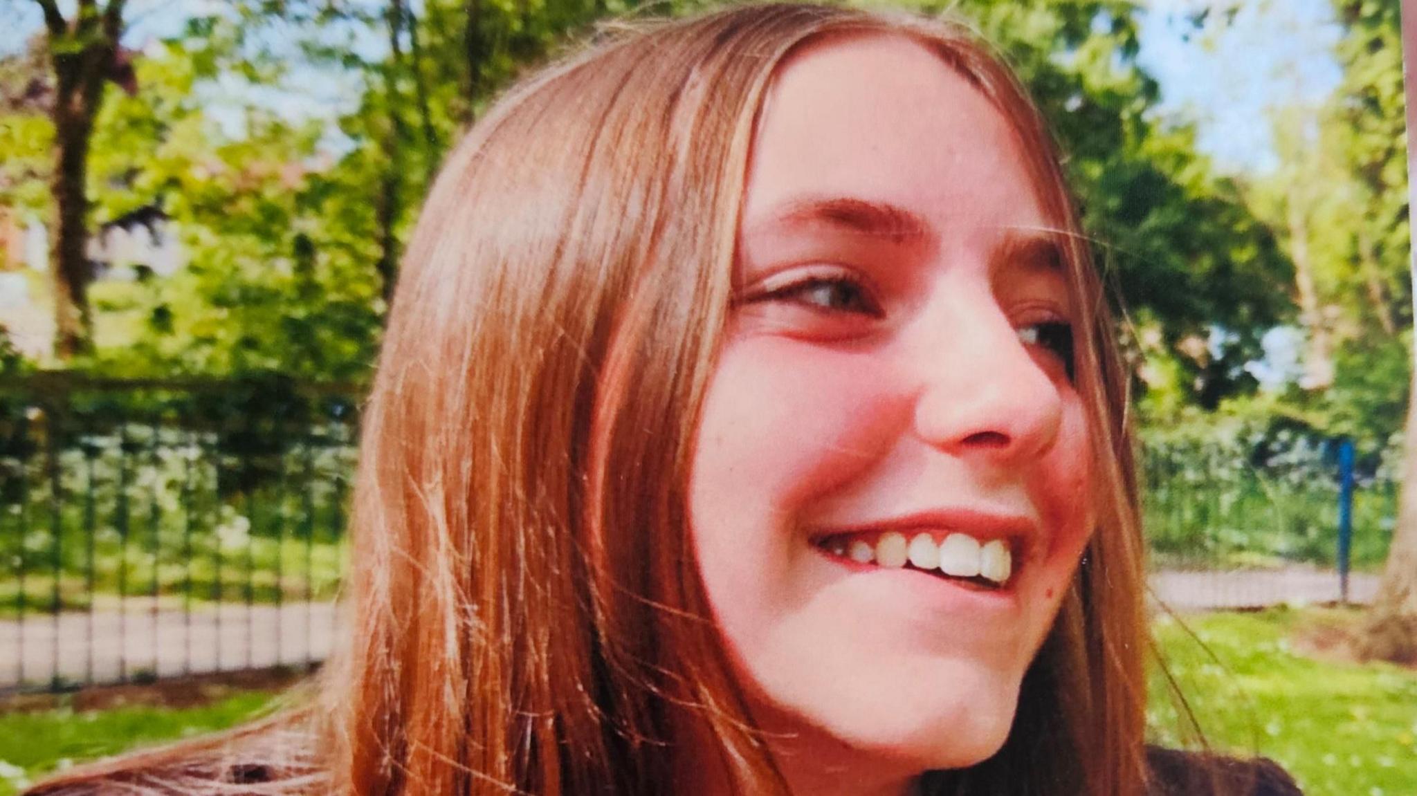 Chloe Longster wearing her high school uniform in a park. She is smiling and her head is turned to the side, she is looking to someone else out of frame to the side. She has long, straight brown hair and it is a sunny day