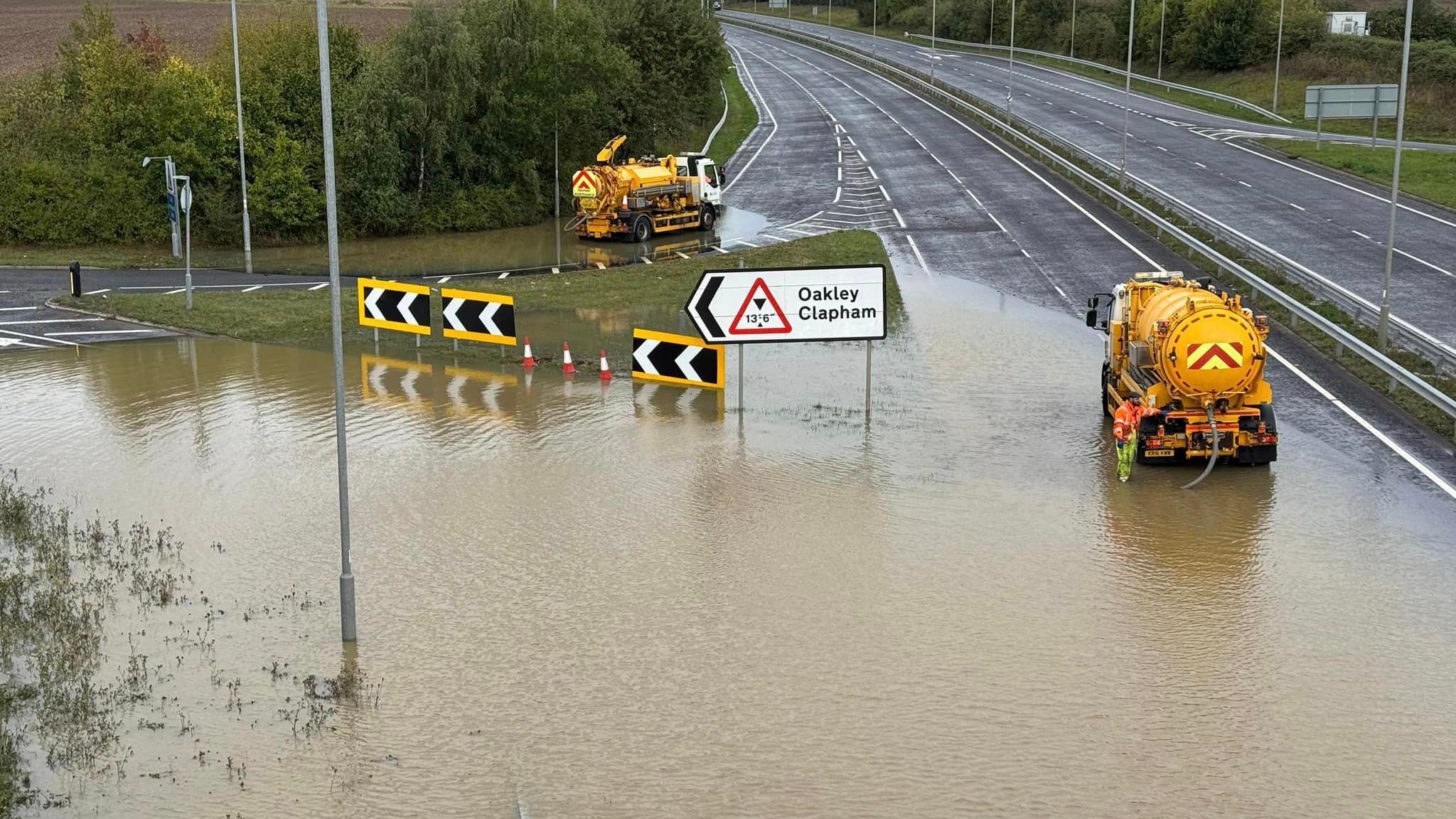 Yellow tank vehicles pump water out of a flooded road junction with a sign saying Oakley Clapham