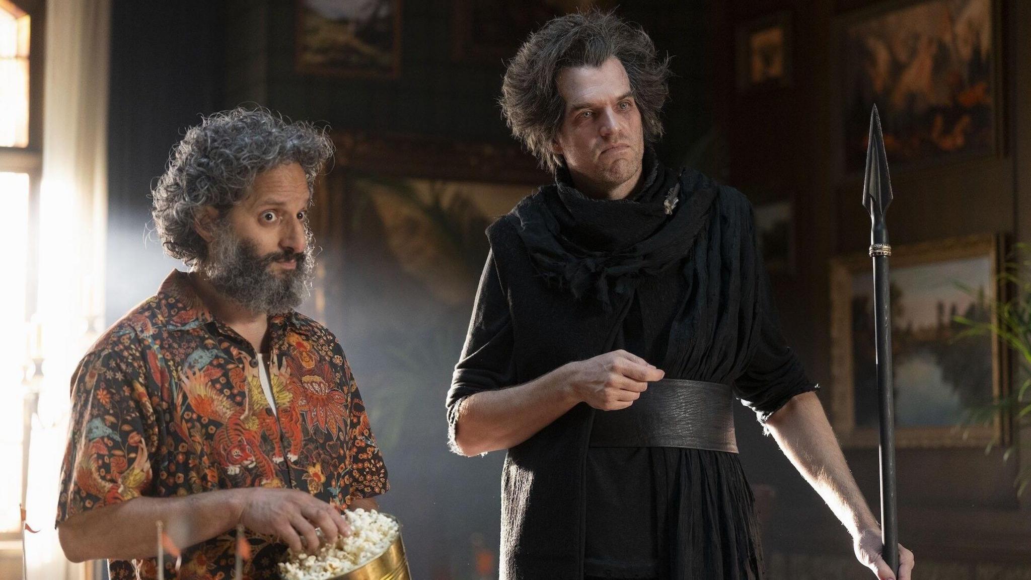 Jason Mantzoukas wears a brightly coloured orange shirt and eats popcorn out of a golden bucket. Timothy Simons dressed in a black robe holding a spear stands next to him.