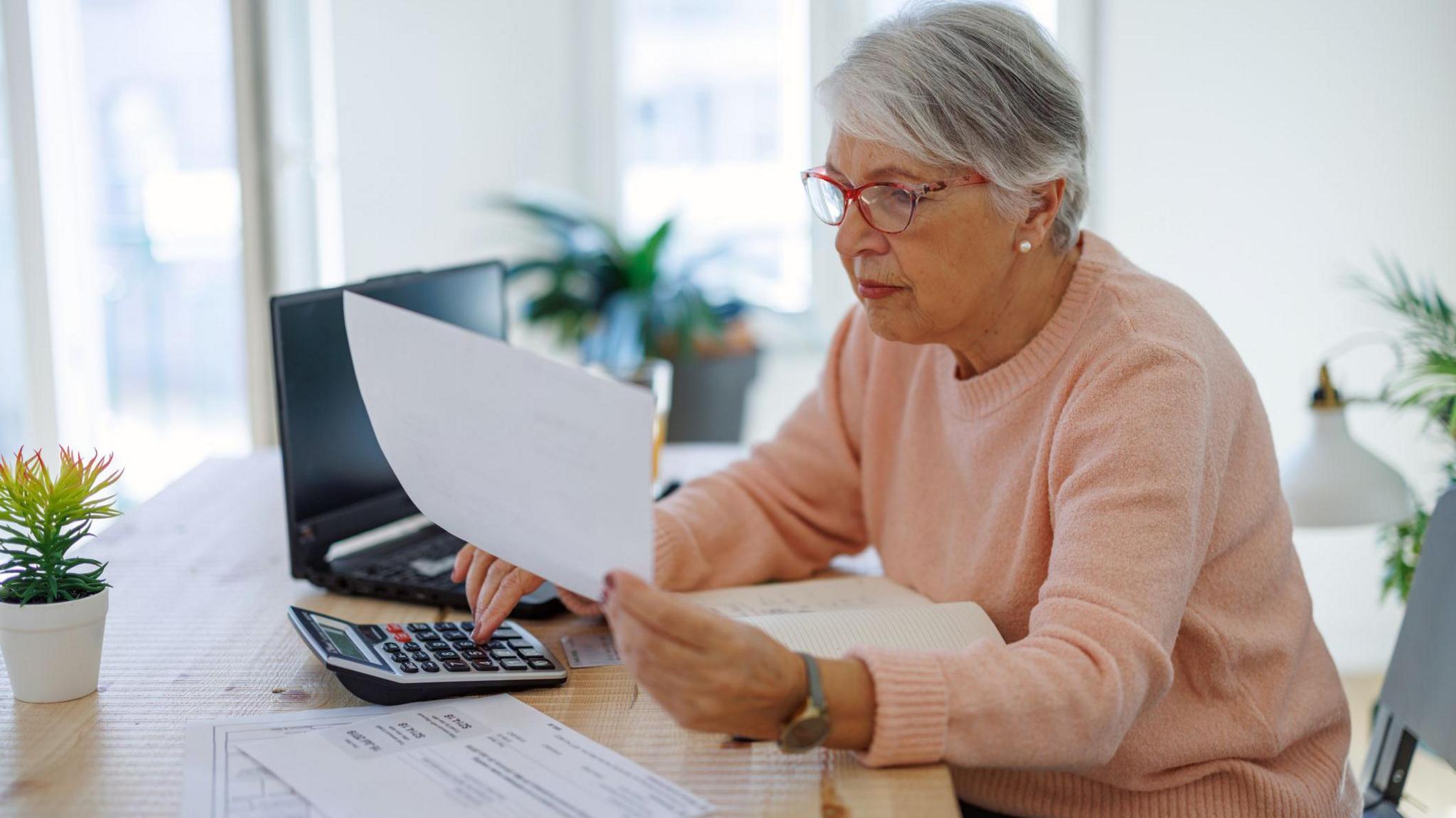 Older woman sits at a desk at home holding a bill with her other hand on a calculator and a laptop in the background.