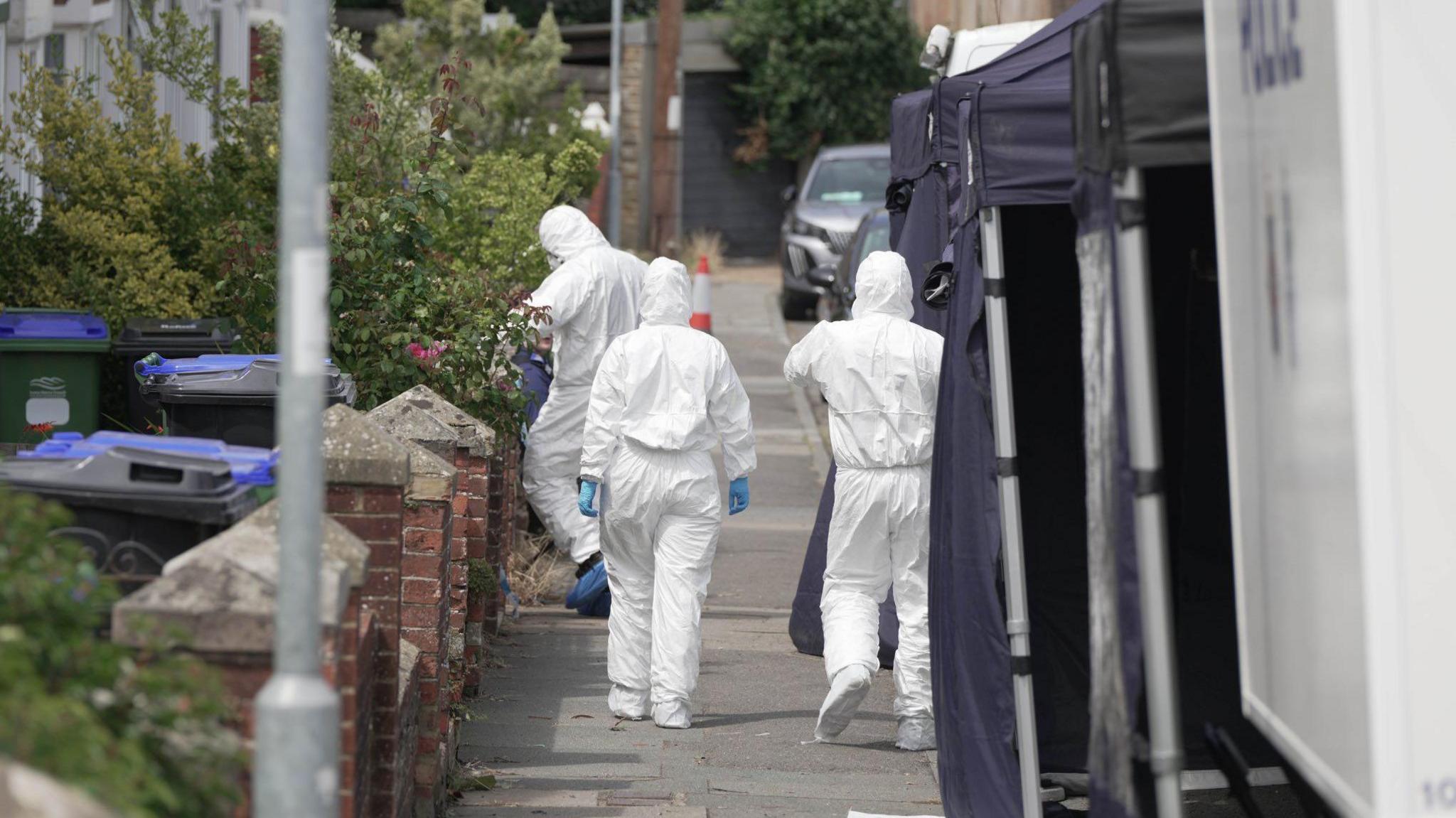 Three people dressed in white forensic bodysuits and protective gloves and shoe covers walk to a terraced property in Newfield Road with pop up police tents in the foreground.