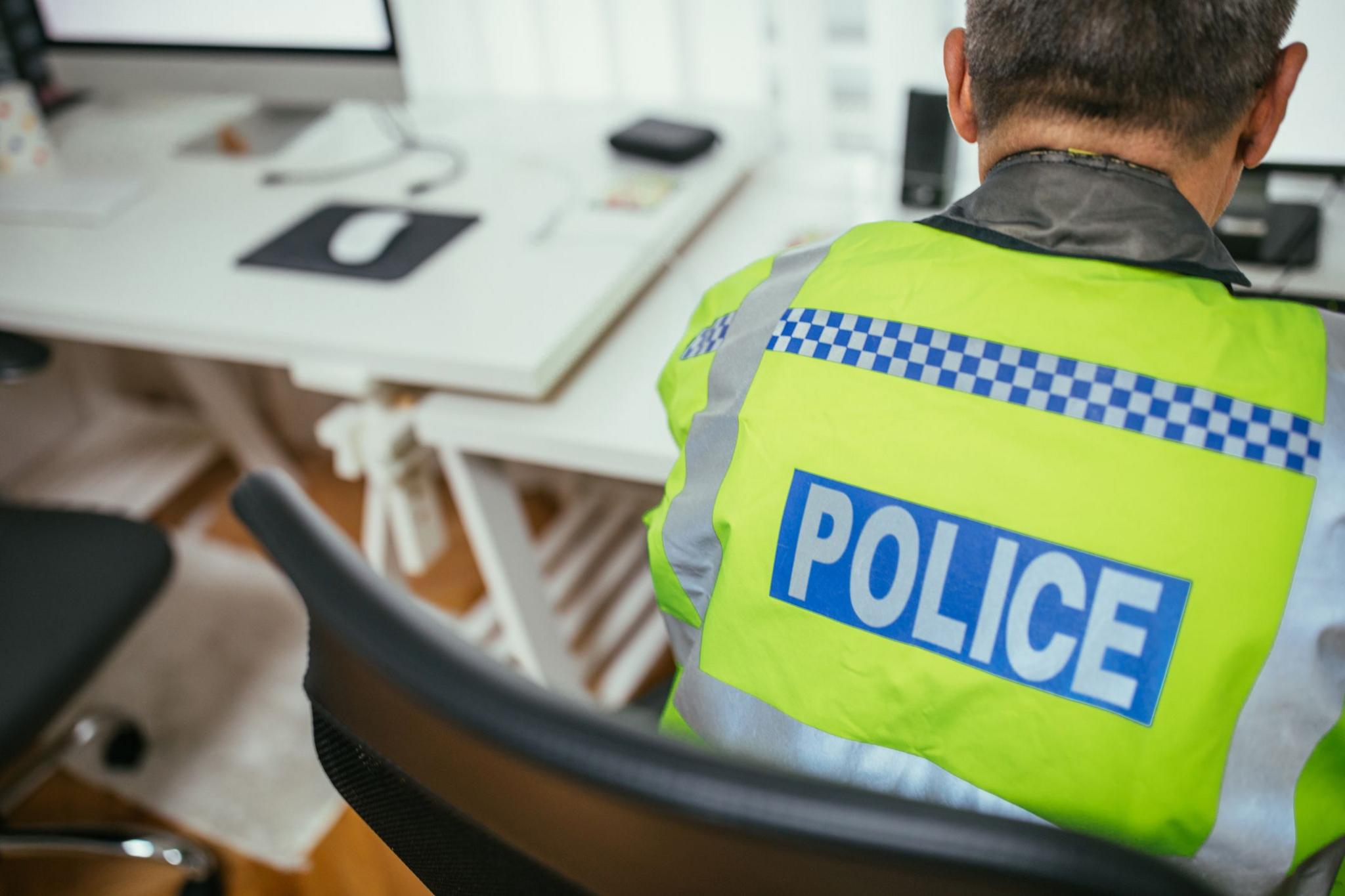 View from rear of a male police officer sitting at a desk, wearing a hi-viz jacket with 'police' written on the back. 