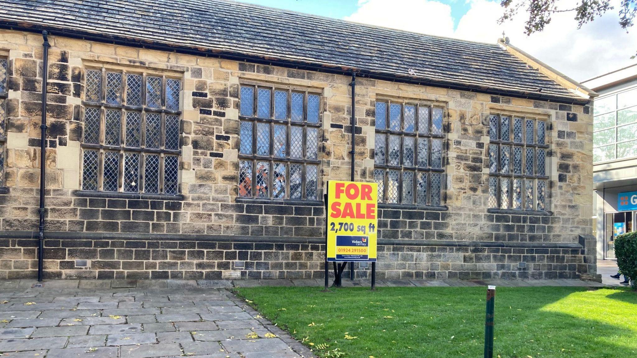 The image depicts a traditional stone building with a slate roof and large rectangular windows arranged in a grid-like pattern. The architecture suggests that the building is quite old, possibly historic. In front of the building, there is a paved walkway that leads up to the entrance, bordered on one side by a neatly maintained grassy area.
Prominently placed on the grass is a bright yellow sign with red text that reads "FOR SALE" in large letters. Beneath that, the sign indicates the property size as "2,700 sq ft." Additional text at the bottom of the sign provides contact details for a real estate agent. The sky overhead is partly cloudy, with some blue sky visible through the clouds.
