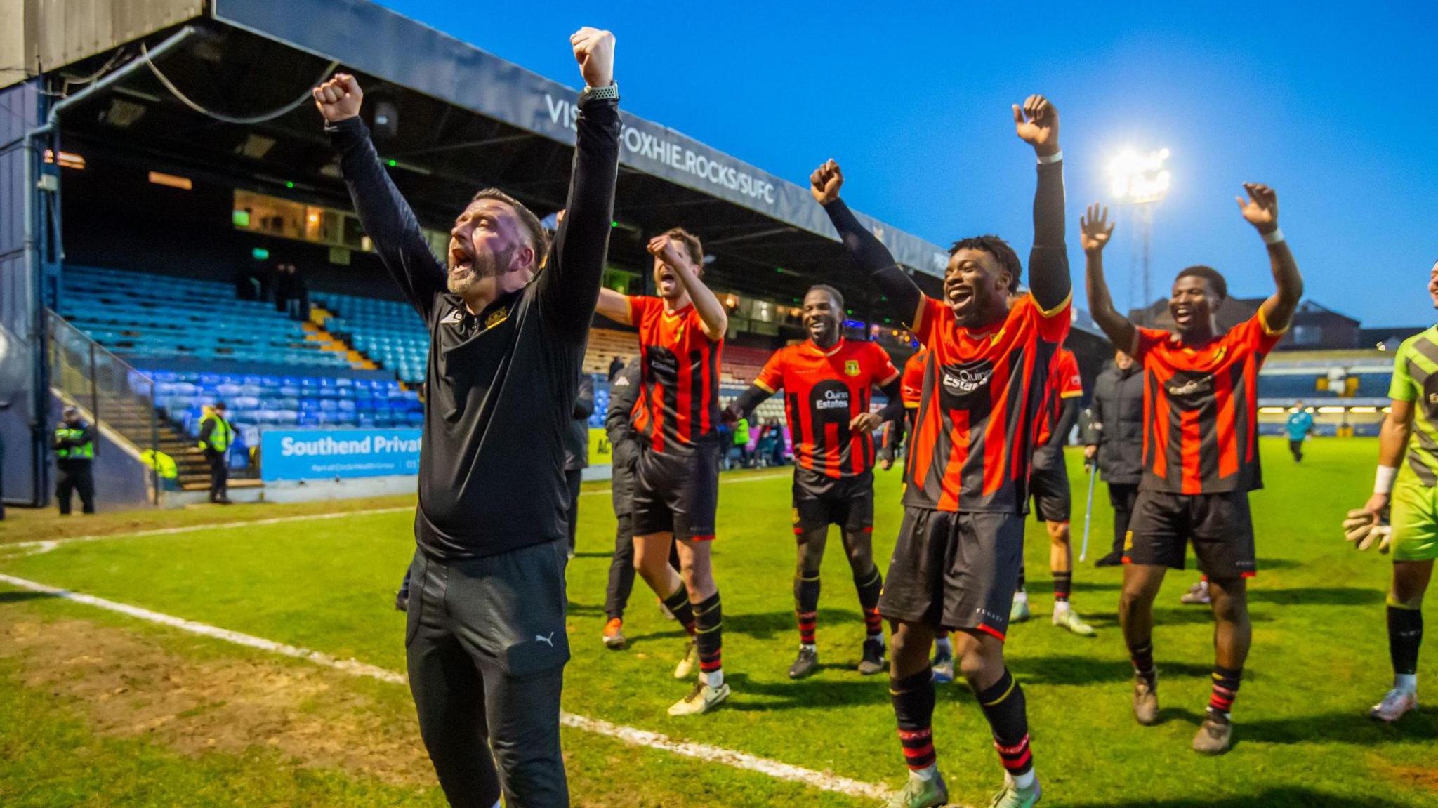 The manager wearing black stands at the forefront of football players who are wearing red and black outfits and cheering