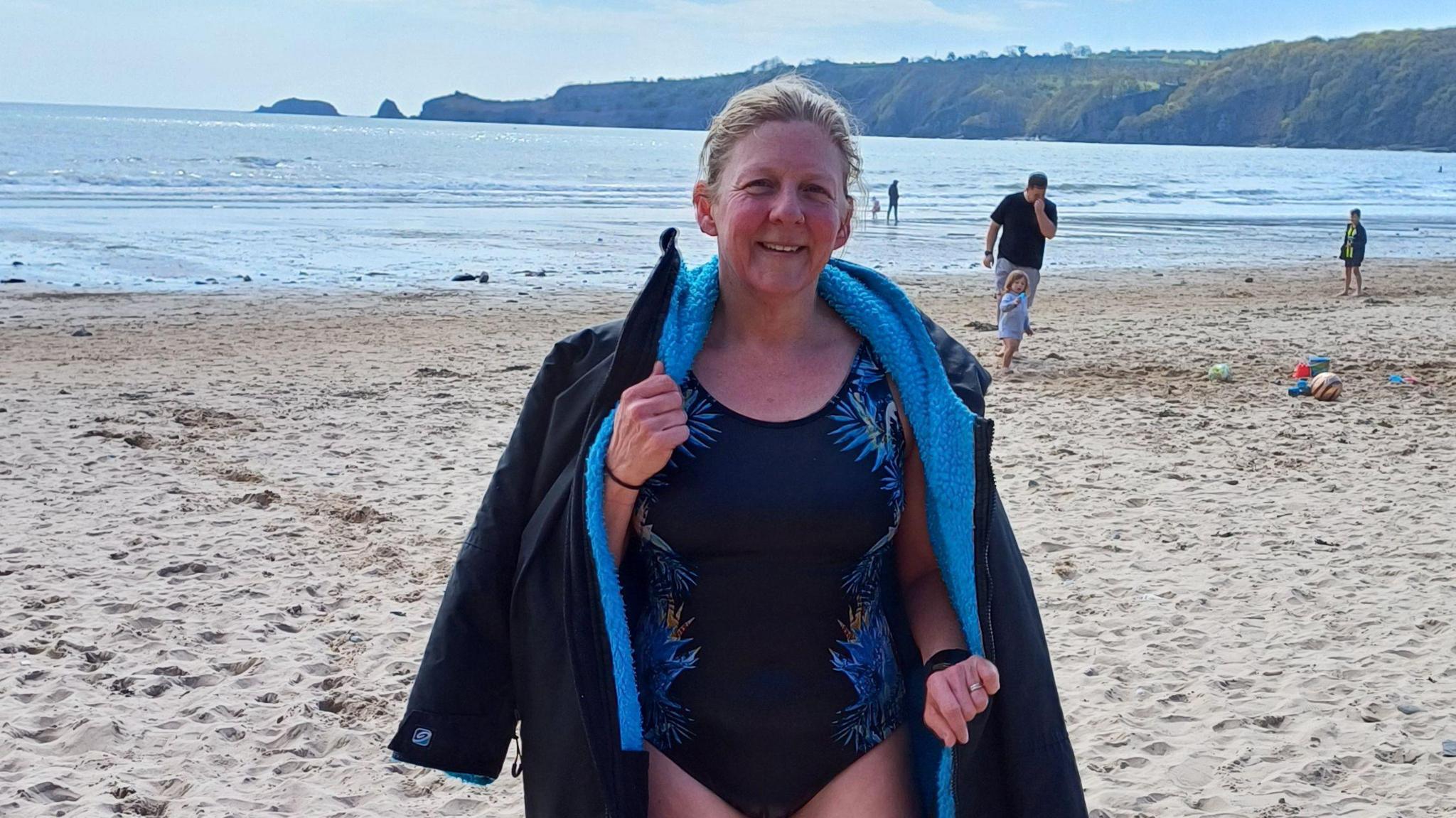 Helene Jewell in her swimming costume and black and blue dry robe at the beach after a swim. You can see the sea and the coastal edge behind her.
