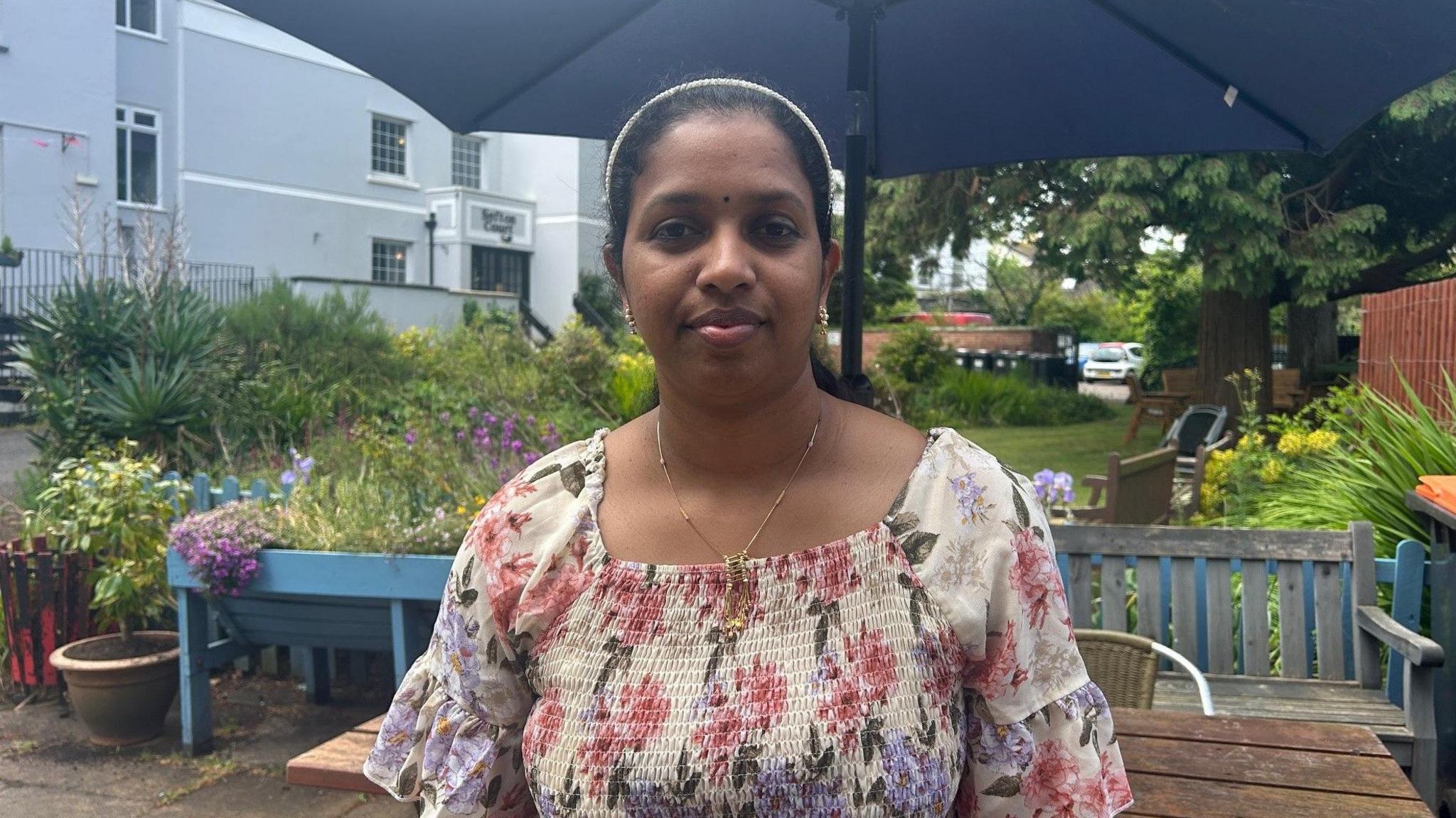 An Asian woman with a floral pink and purple dress standing under a large patio umbrella