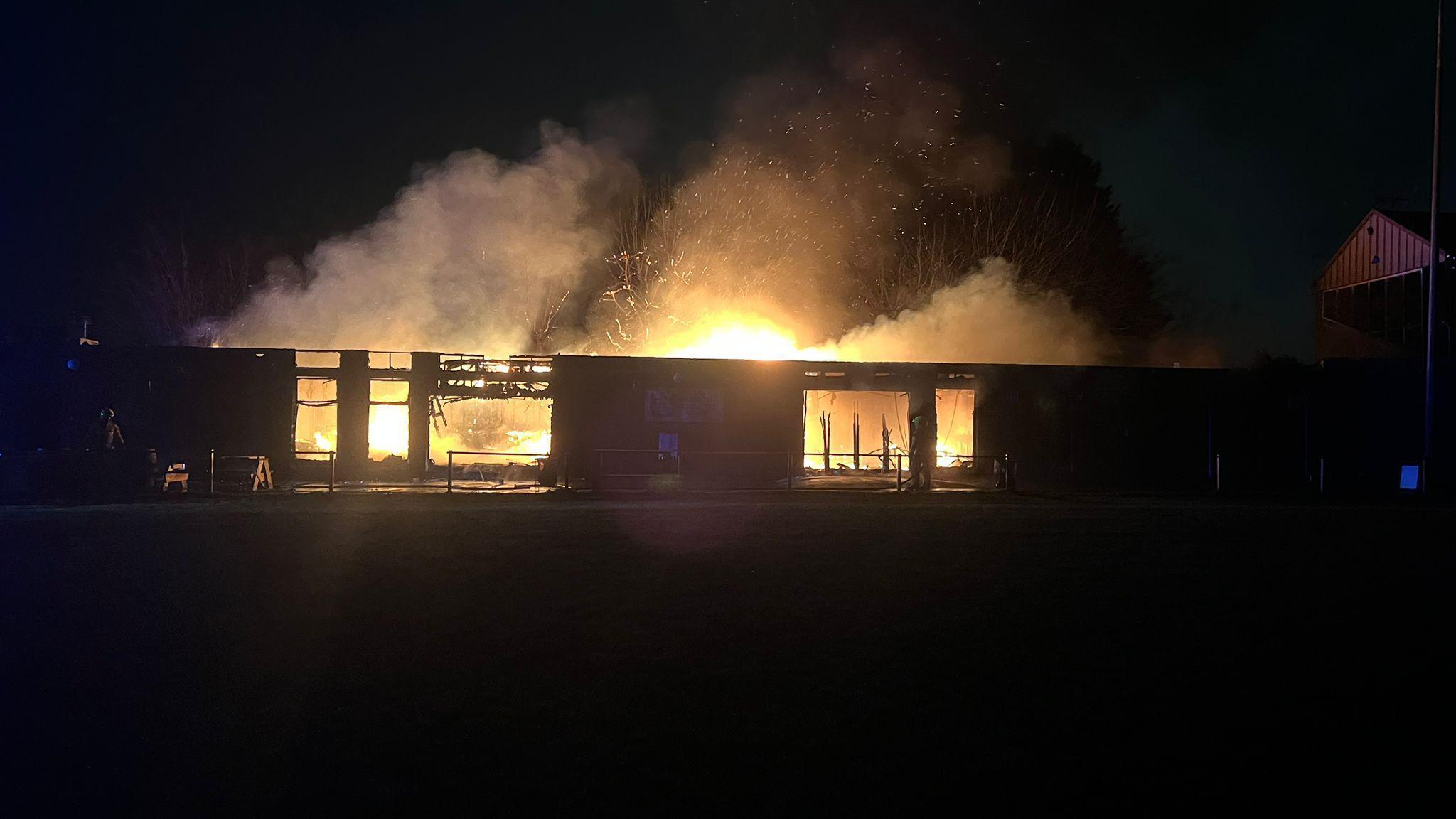 A low-rise building on fire; it is night-time and flames and smoke can be seen billowing from the clubhouse