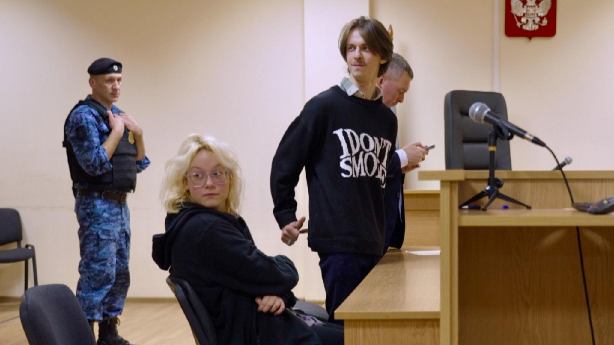 Diana sits in court while Alexander stands. A man in a blue combat uniform and a flak jacket looks on.
