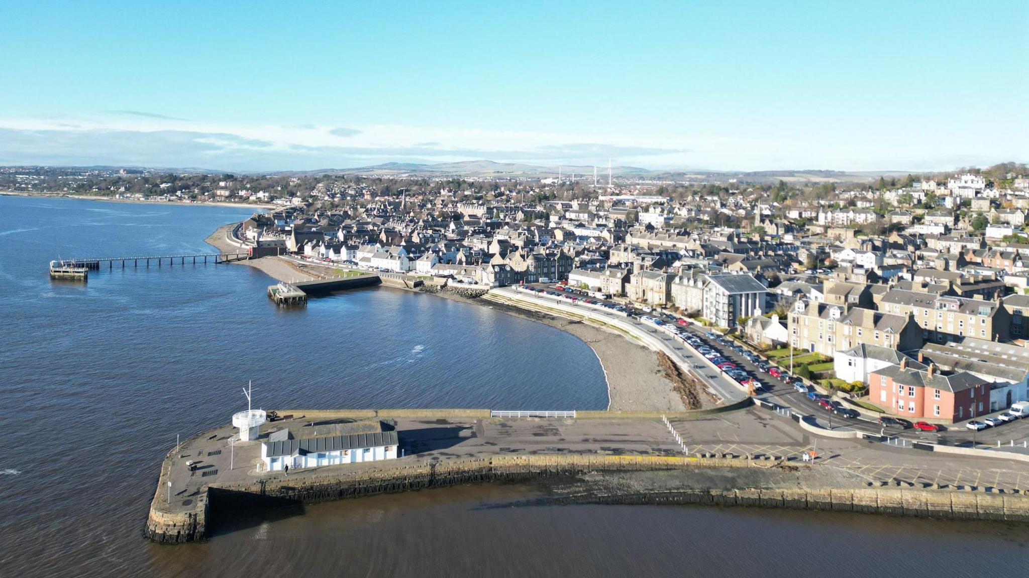 An view of the seaside town of Broughty Ferry. The sea is on the left side of the picture, and the town is on the right, with hills visible in the distance. In the foreground, a pier stretches out into the water.
